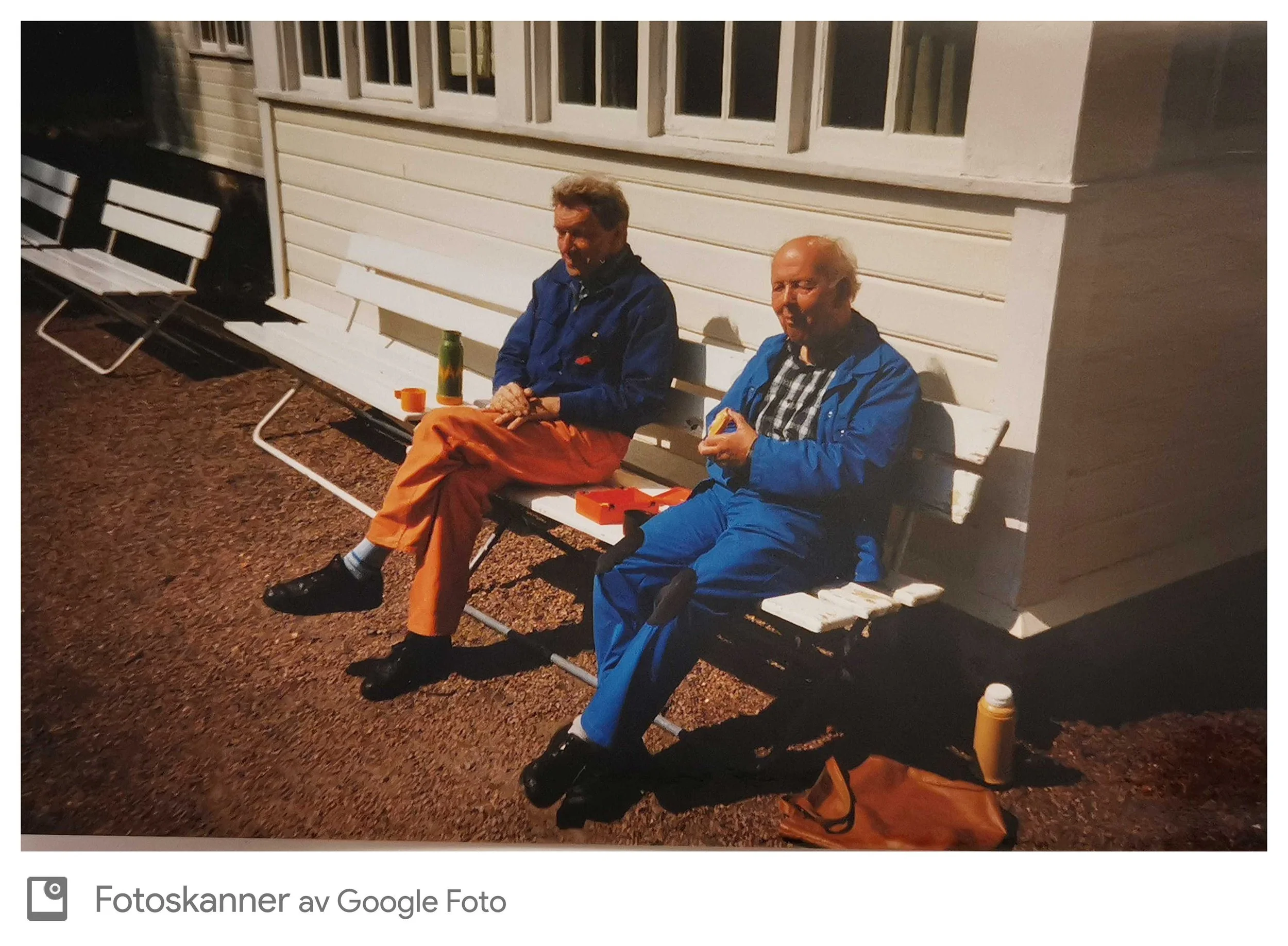 To eldre menn sitter på en hvit benk utenfor en bygning om kvelden. Den ene har blå jakke og oransje bukser, den andre har blå jakke og blå bukser. Begge har noe å spise. Det er noen ting på bakken foran dem, inkludert en oransje veske og en flaske.