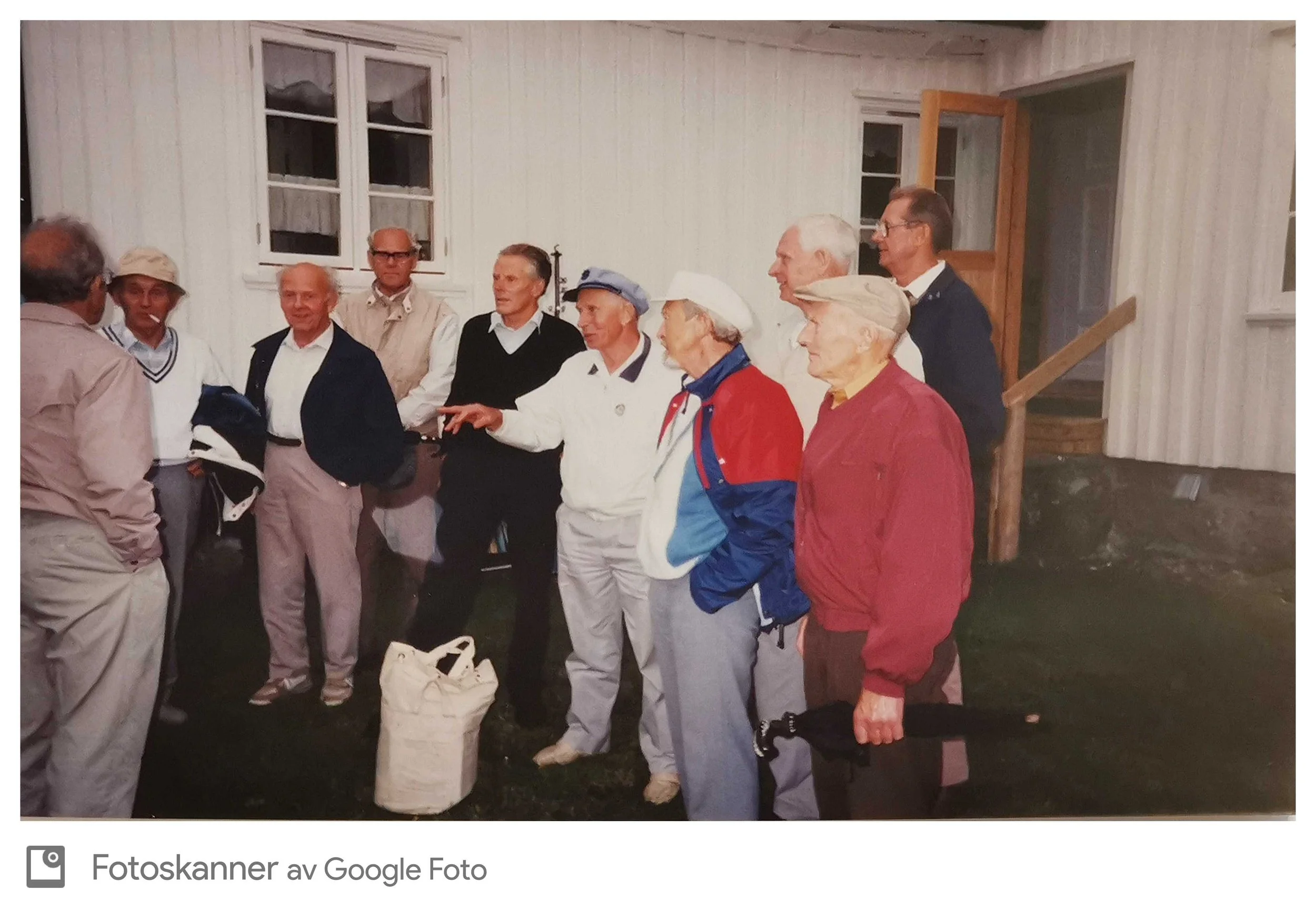 En gruppe eldre mennesker står sammen utendørs og prater. De har på seg komfortable klær, noen har luer og solbriller. Det ser ut til å være kveldstid. Det er en hvit husvegg og et vindu i bakgrunnen.