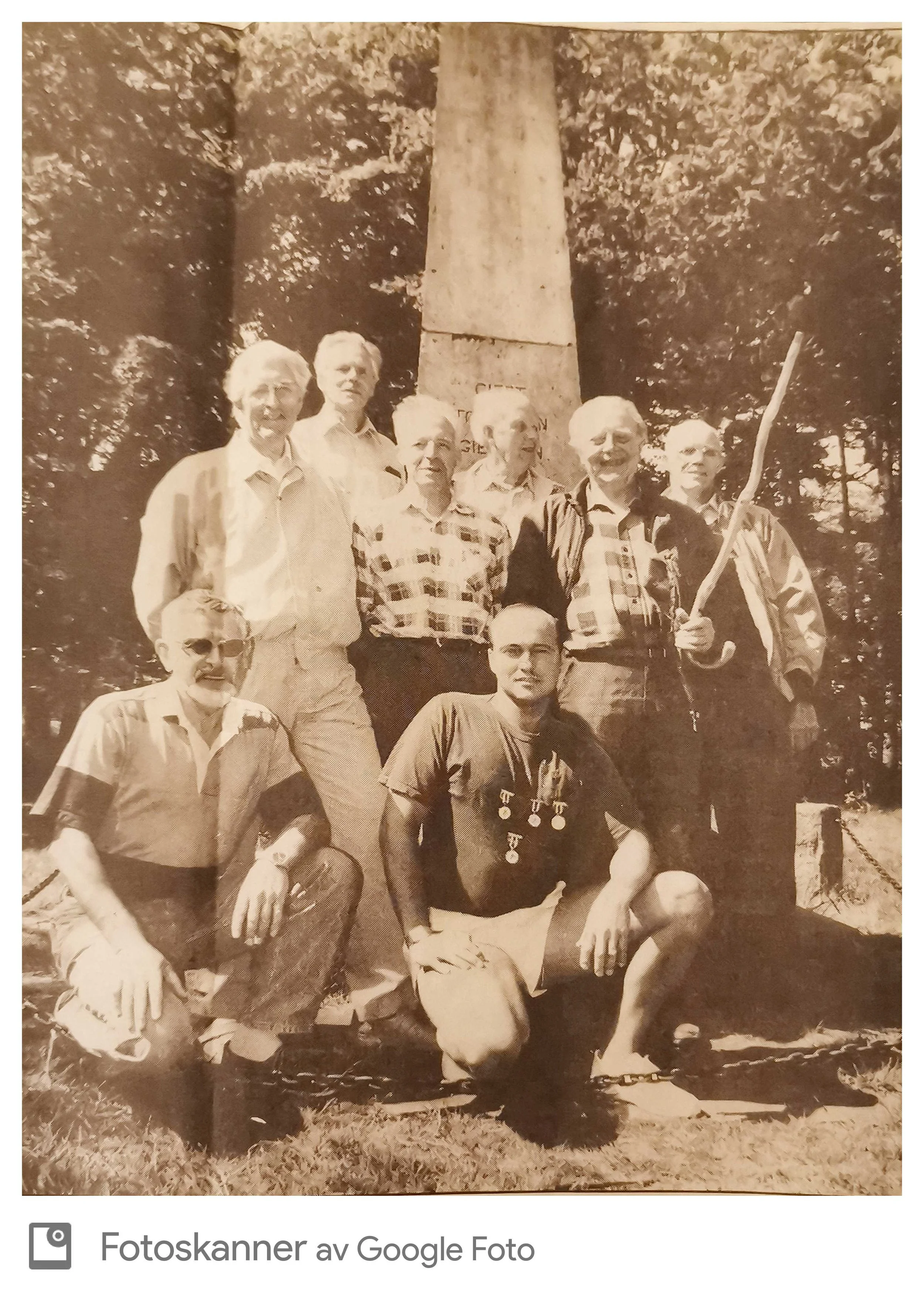 Gruppe eldre mennesker som poserer foran en steinmonument, noen av dem har medaljer rundt halsen, og en person holder en pinne. Det er trær i bakgrunnen.