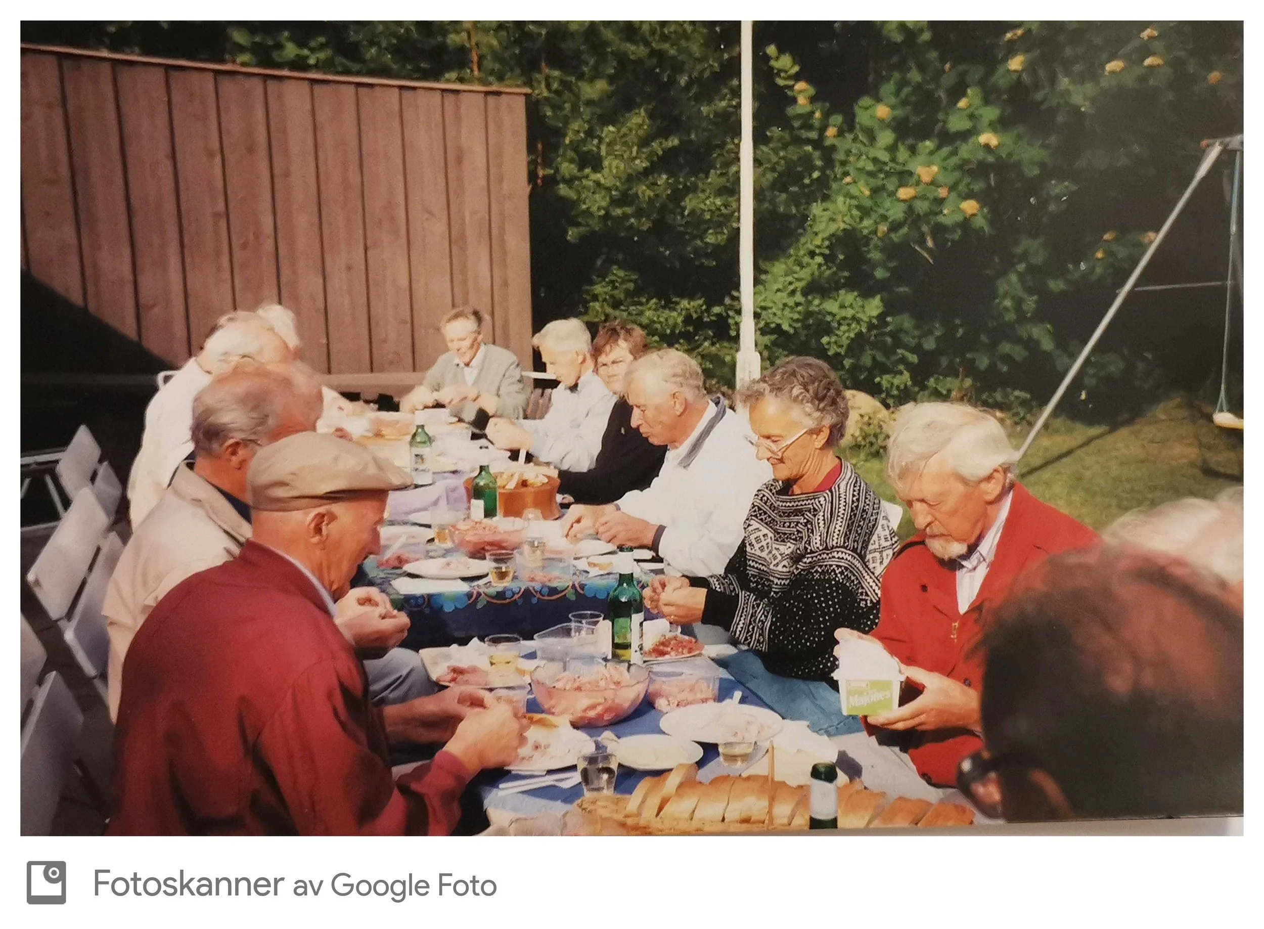 Grupp mennesker som spiser ute ved et langbord på en utendørs brunsj eller fest, omgitt av grønne trær og kveldshimmel.
