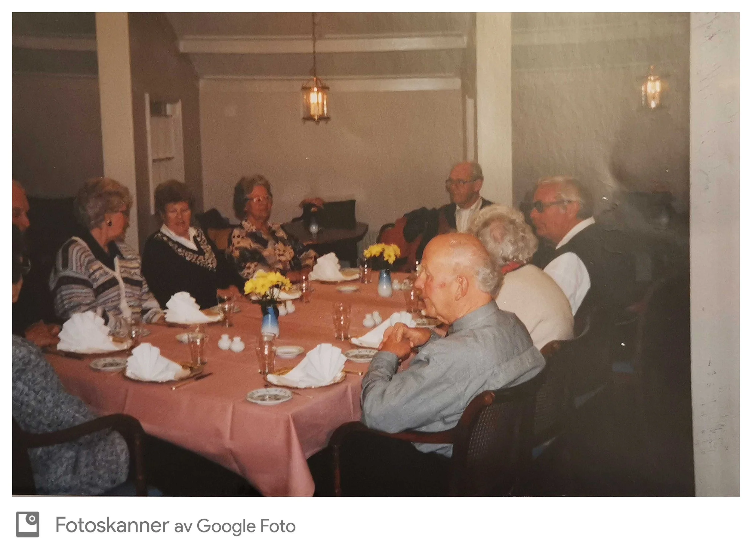 Et bord dekket med konserver og blomster, der mennesker sitter rundt og snakker.