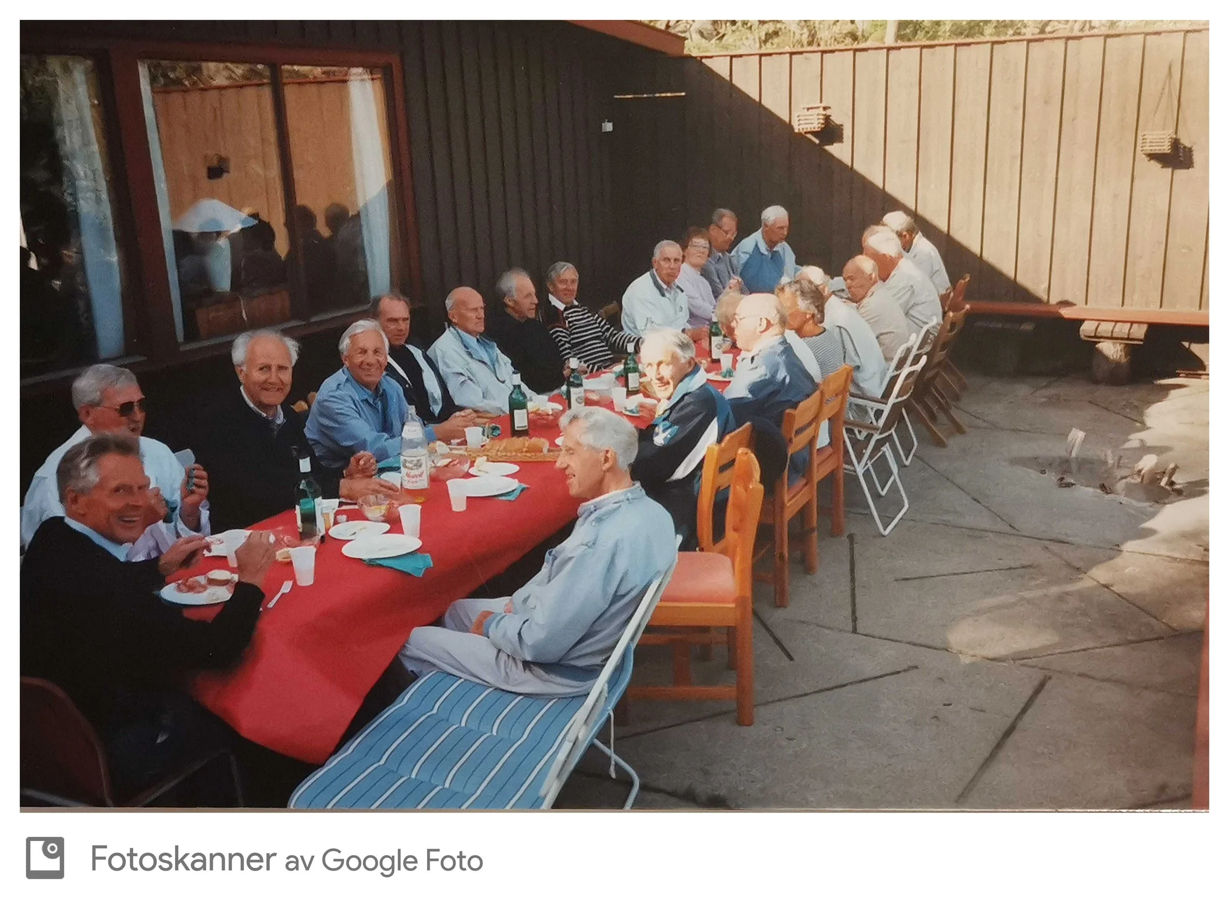 En gruppe eldre mennesker sitter rundt et langbord med rødt duk og nyter et måltid utendørs. De ser glade ut og har på seg casual klær.