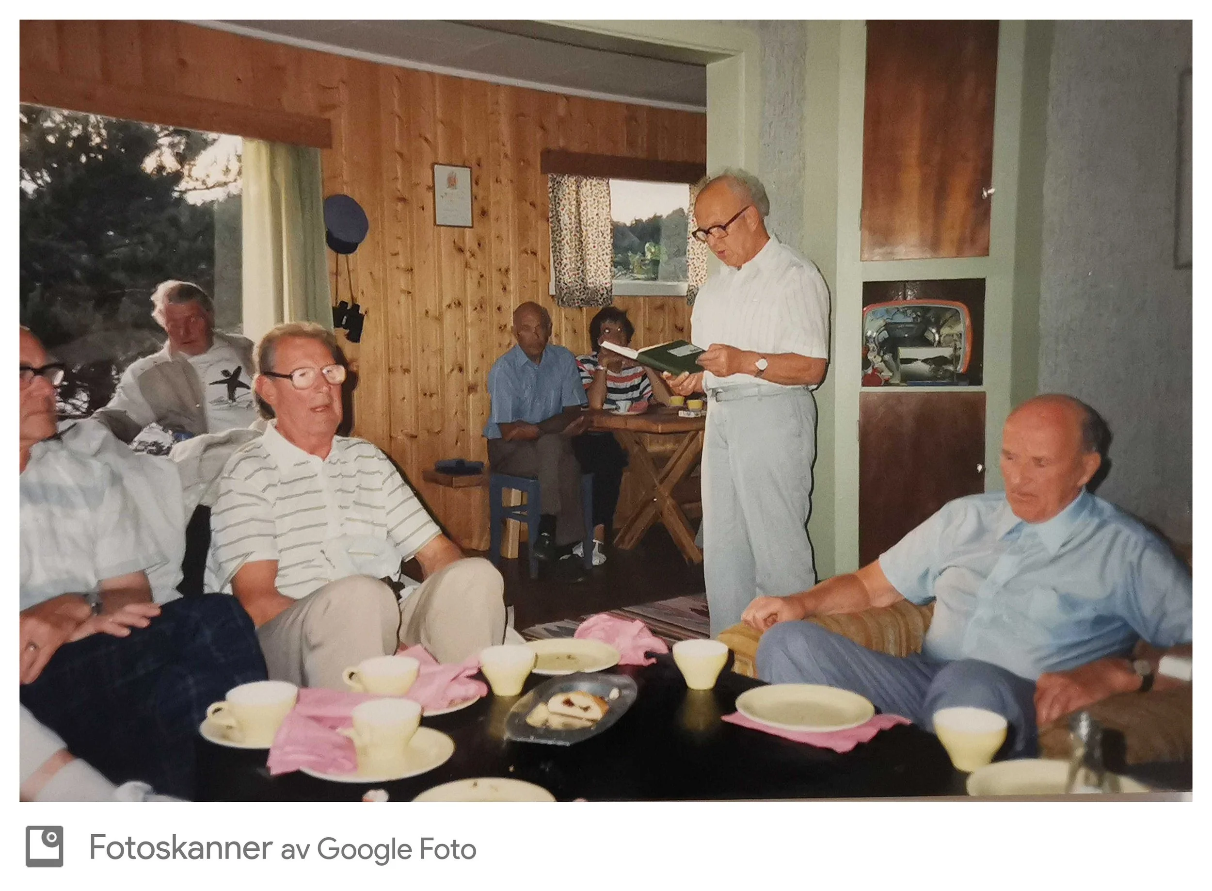 Interiørbilde av en gruppe mennesker som deltar i en sammenkomst eller møte i et rom med trepanelvegger. Flere personer sitter rundt et bord med papptallerkener og mat, mens en eldre mann står og leser fra en bok. Det er vinduer med blomstrete gardin