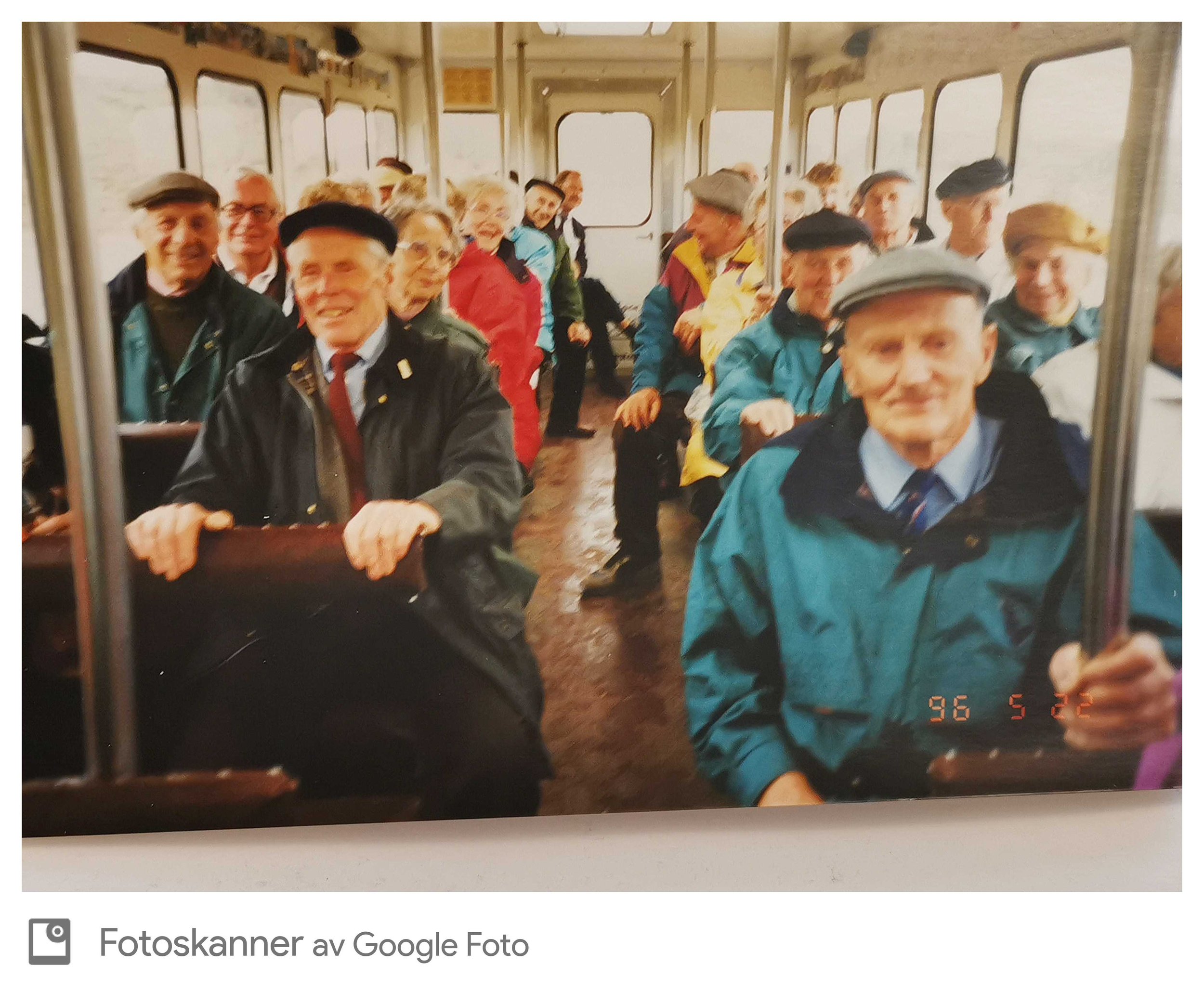 En gruppe eldre mennesker smiling og som sitter i en vogn eller kjørerjern på en vogn, inne i en trikk, med store vinduer rundt. Noen av dem har på seg capser og regnjakker. De ser ut til å ha det moro og nyte turen.