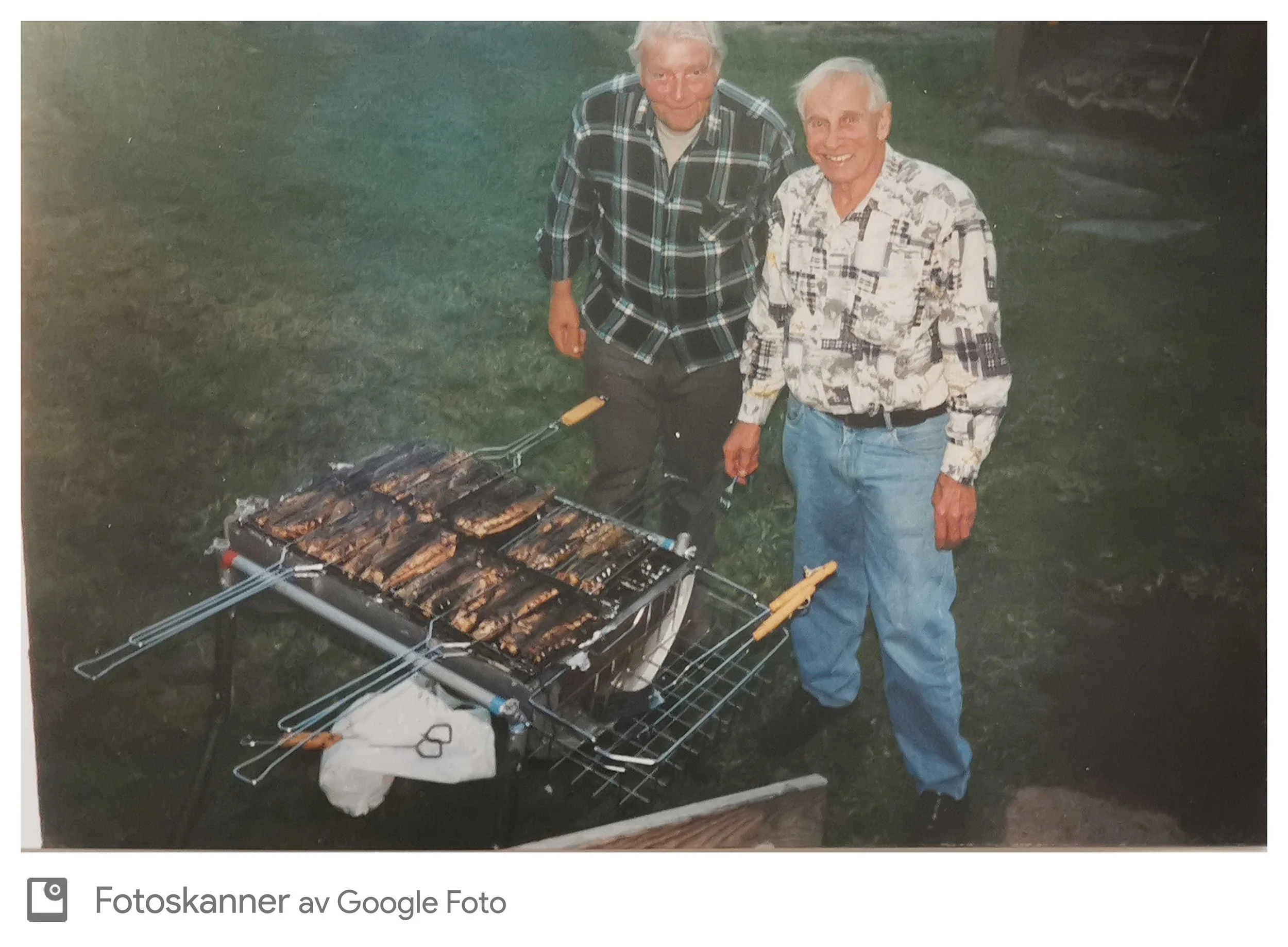 To eldre menn står ved en grill med fisk, i en grønn hage. De smiler og ser fornøyde ut.