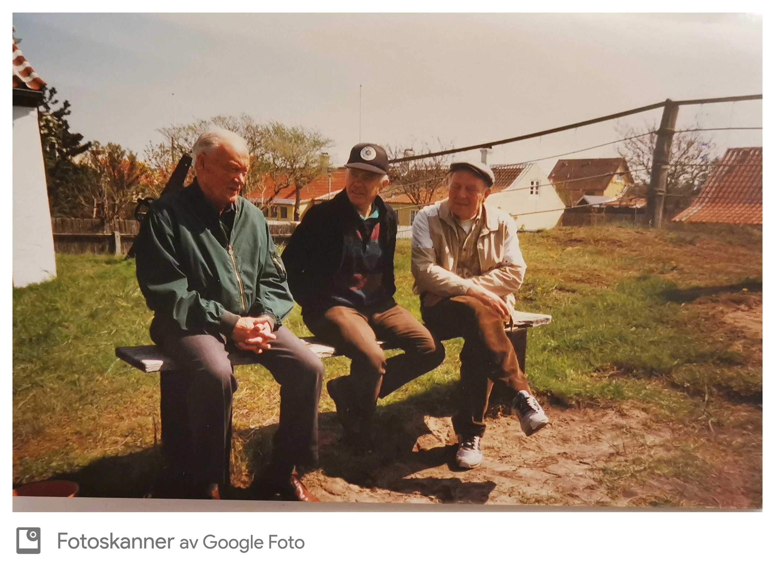 Tre menn som sitter på en benk i en hage med hus i bakgrunnen.