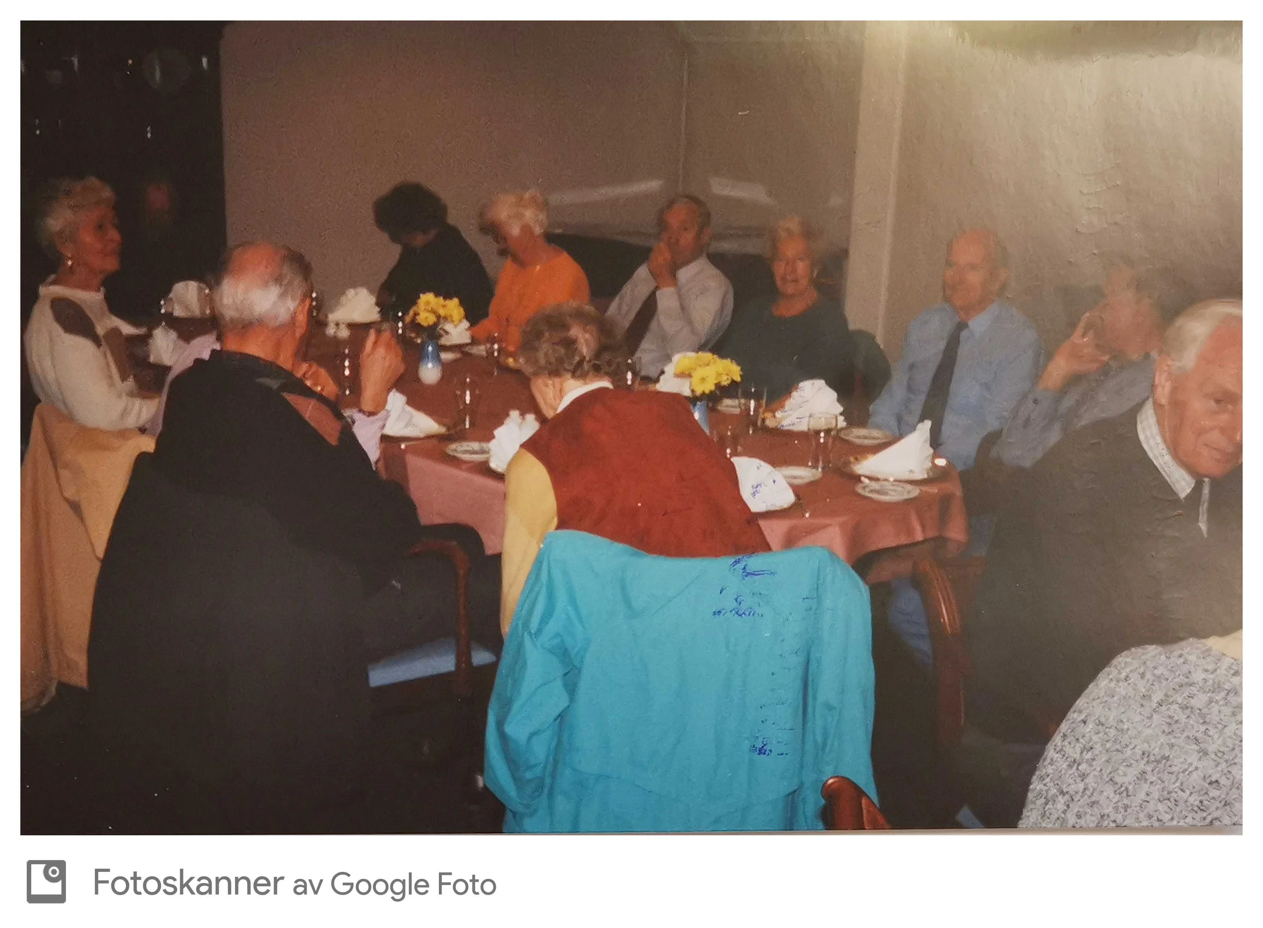 Familie eller gruppe mennesker sitter rundt et langbord under en sammenkomst, med blomsterdekorasjoner og mat på bordet, i et rom med lyst trepanel og malte vegger.