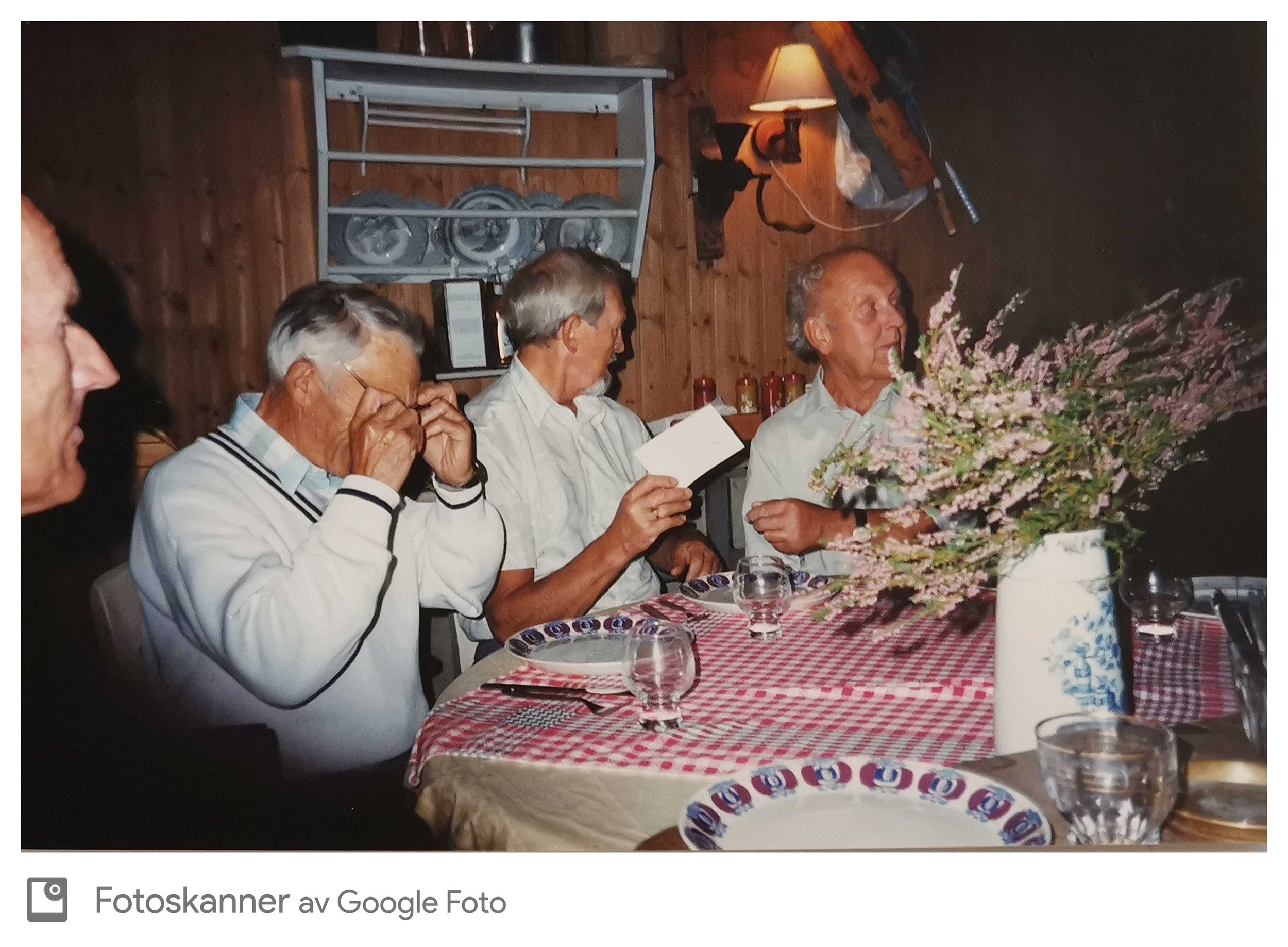 Aksjon på en restaurant eller et festlokale med eldre menn sittende ved et bord dekket med et rødt og hvitt rutete duk. Et stort blomsterarrangement og flere Dr. Oetker-produkter er på bordet. Mørk vegg med trepanel og et skap med vektere i bakgrunne