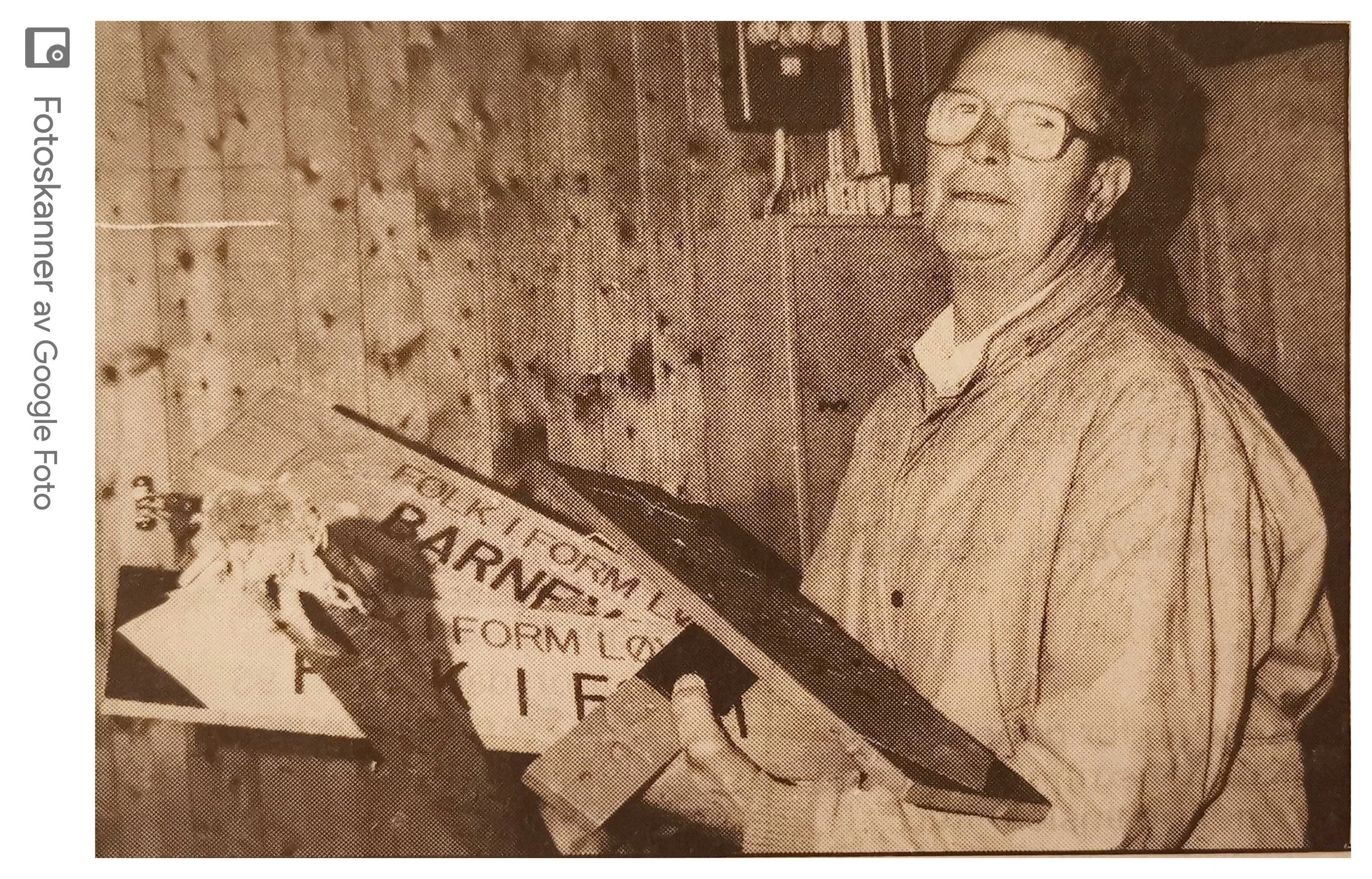 En eldre kvinne med briller og løstsittende hår står foran en vegg med bilder og plakater, holder en bunke aviser i hendene.