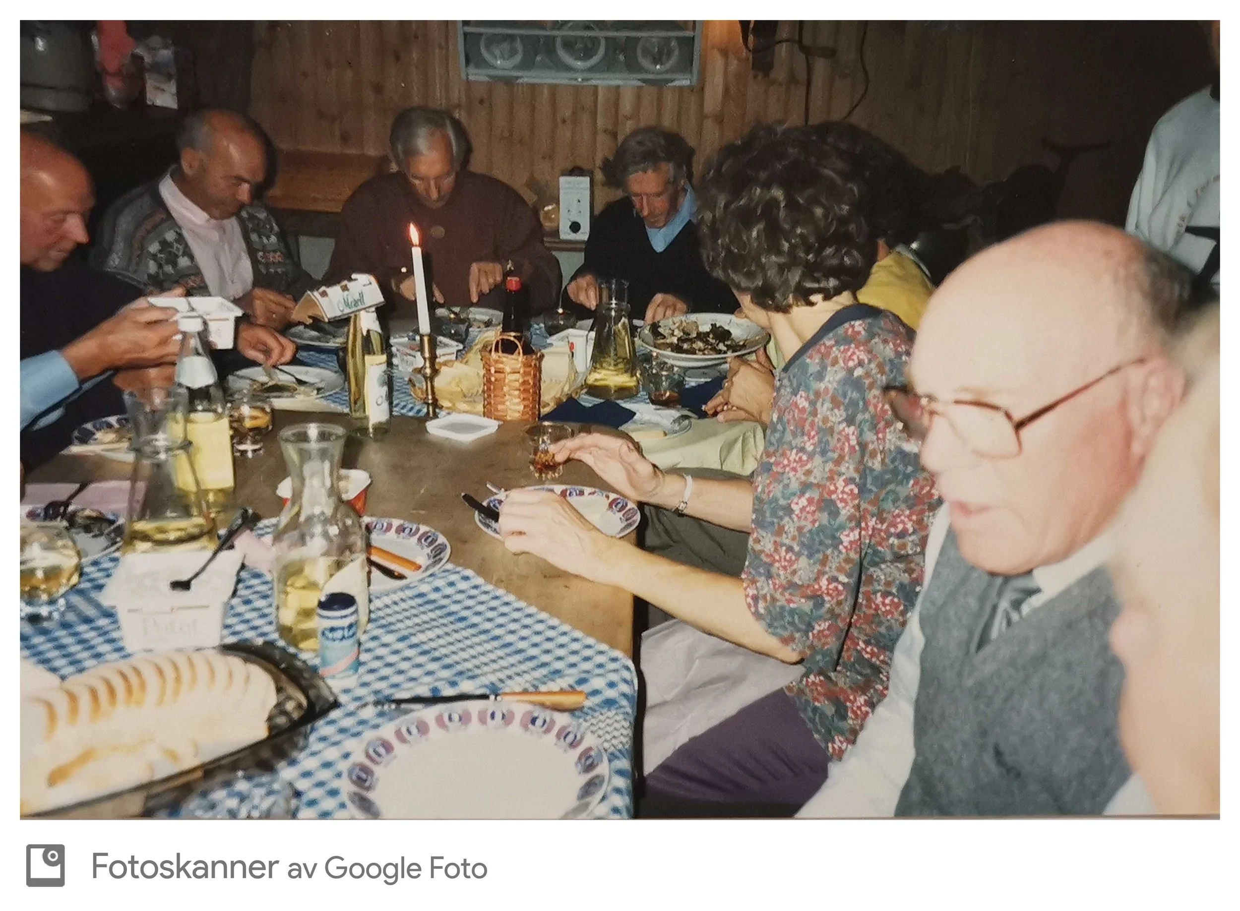 En gruppe mennesker sitter rundt et bord og spiser i et rom med trepanelvegger. Det er en lysende lysestake i midten av bordet, og folk ser ut til å være opptatt av måltidet eller noe på bordet.