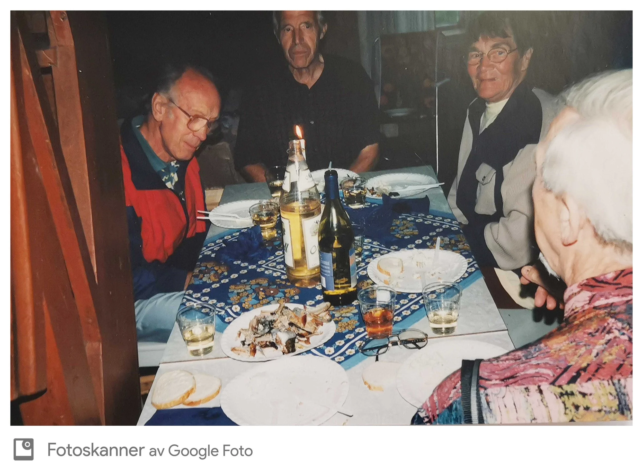 Menn og kvinner rundt et bord med mat og drikke under en sammenkomst, innendørs, med matrester og glass på bordet.