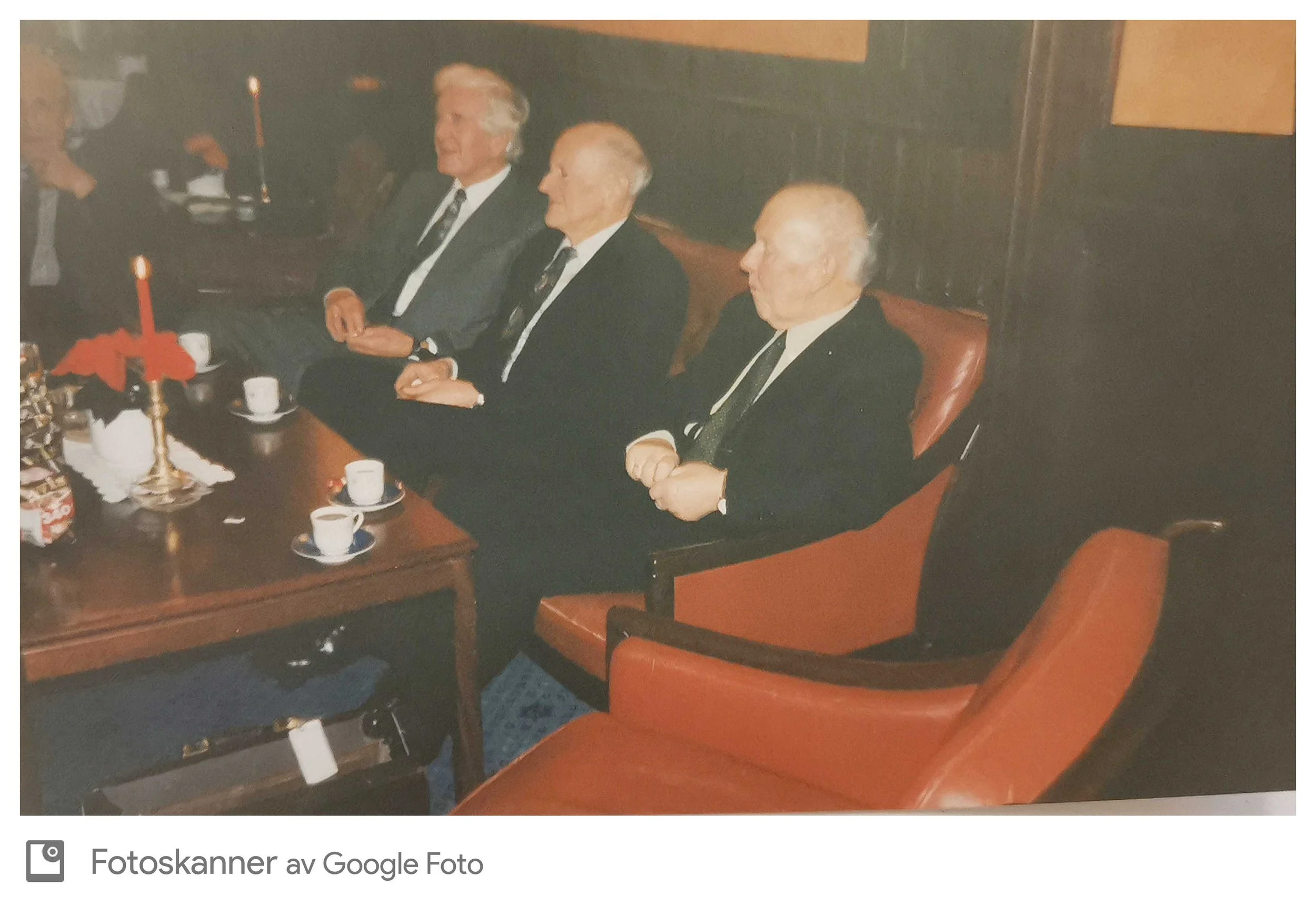 Tre eldre menn sitter i en konferansesal, kledd i dress og slips, og ser ut til å være i en formell sammenkomst.