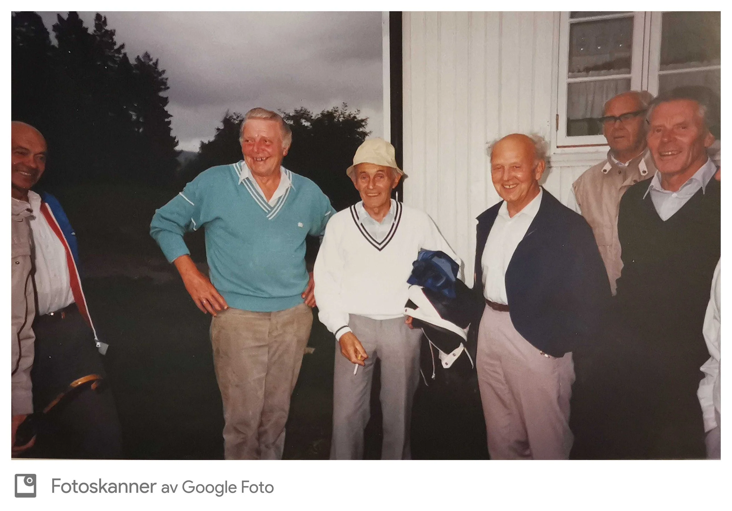Gruppe av fem eldre menn som smiler og står sammen utenfor en bygning, noen med klær som ser ut til å være tilknyttet golf, under en overskyet himmel.
