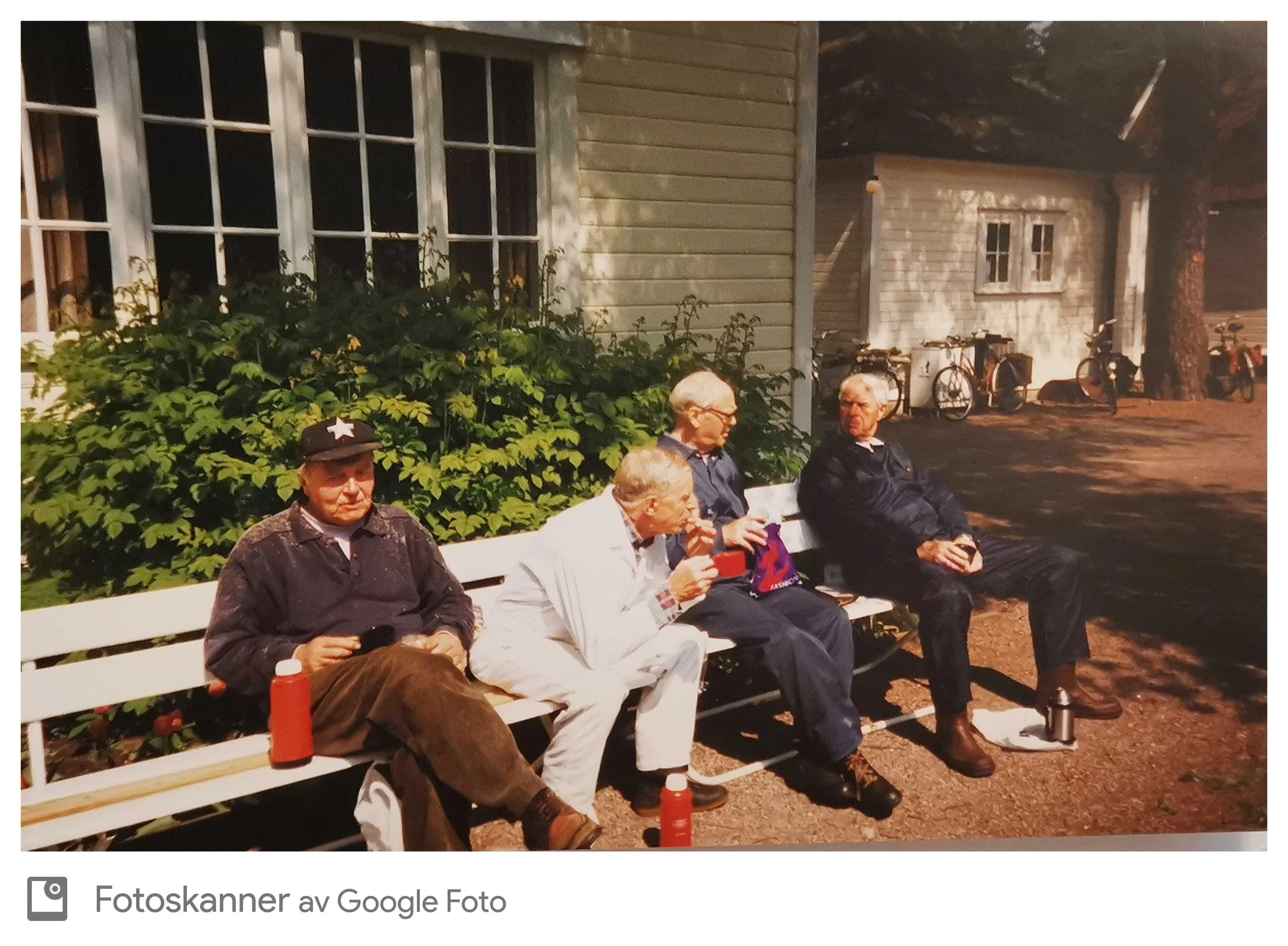 En gruppe eldre mennesker sitter på en parkbenk om kvelden, omgitt av grønnsaker og planter, foran en liten trebygning og flere sykler, i et skogkledt område.