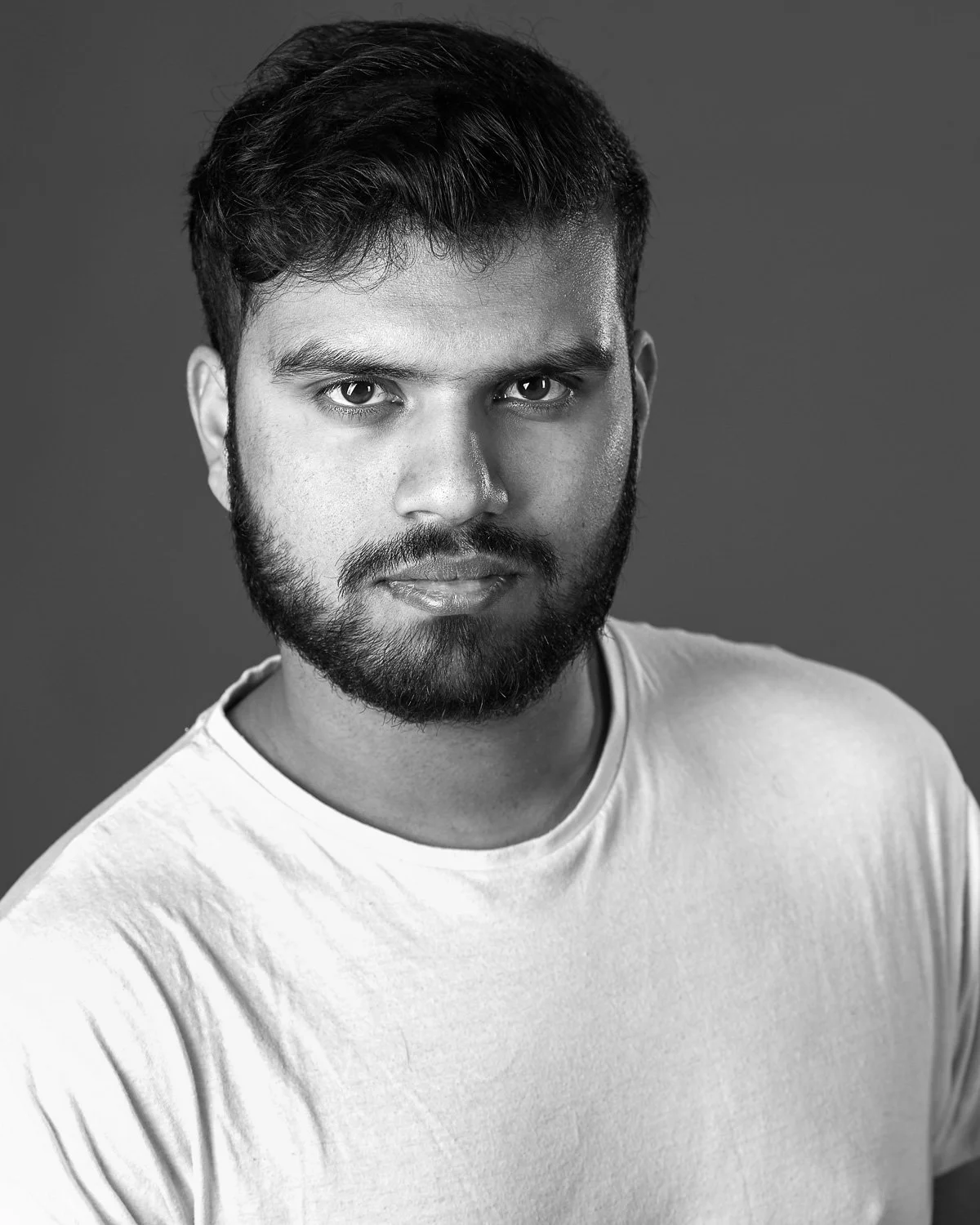 Black and white portrait of a young man with dark hair and a beard, wearing a plain light-colored t-shirt, looking directly at the camera with a neutral expression.