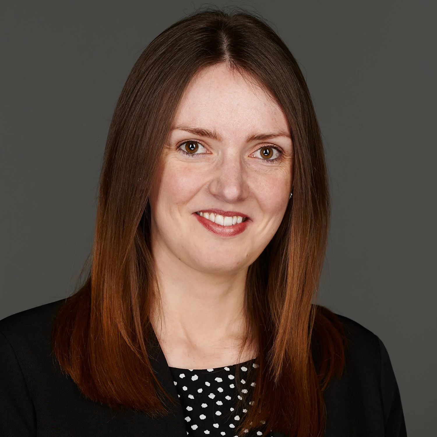 A professional woman with shoulder-length brown hair, wearing a black blazer and polka dot blouse, smiling against a dark gray background.