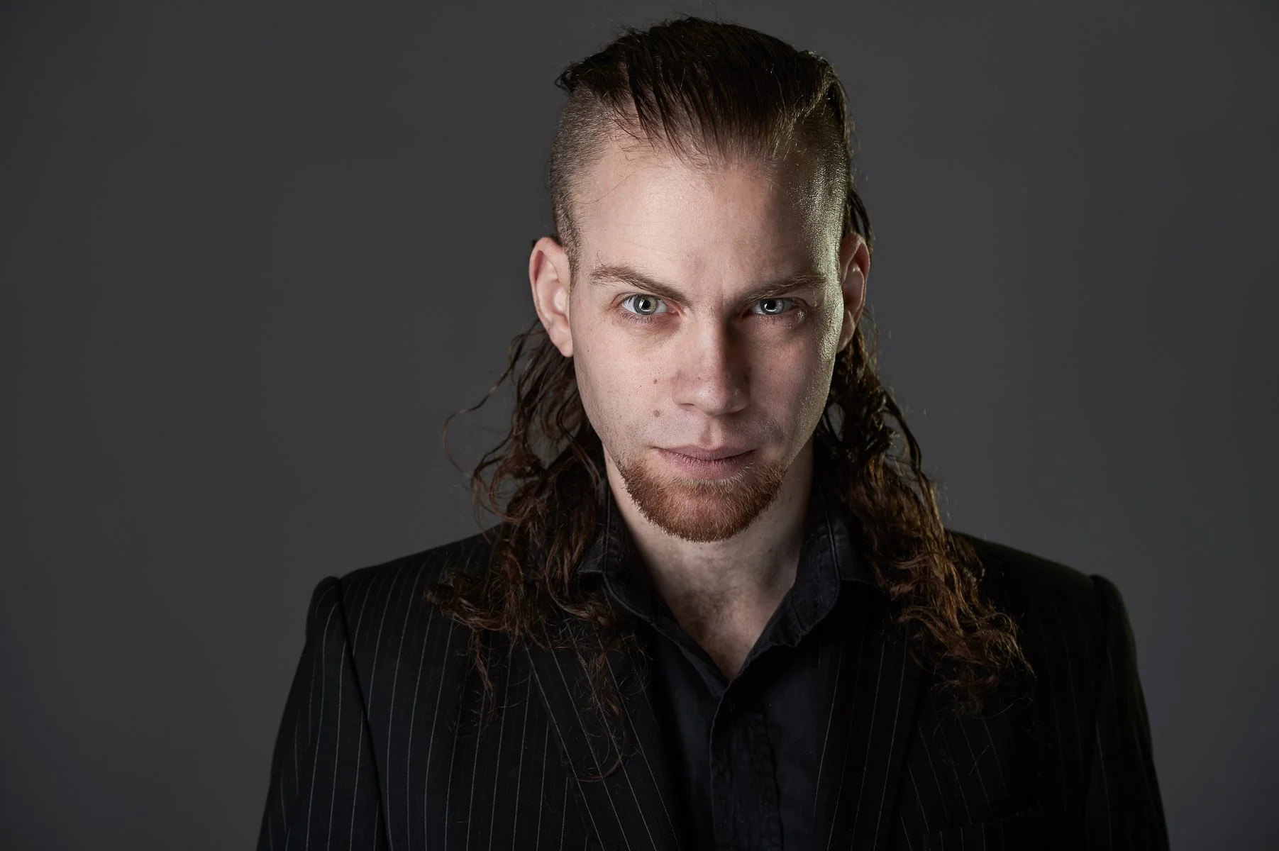 A man with long curly hair, a beard, and blue eyes wearing a black pinstripe suit with a black shirt, looking directly at the camera against a dark gray background.