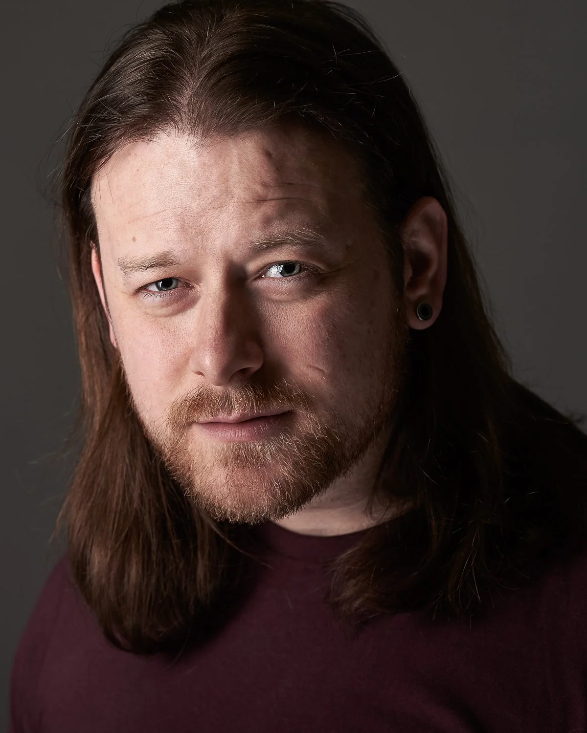 Close-up of an intense man with long brown hair, blue eyes, and a beard, looking at the camera against a dark background.