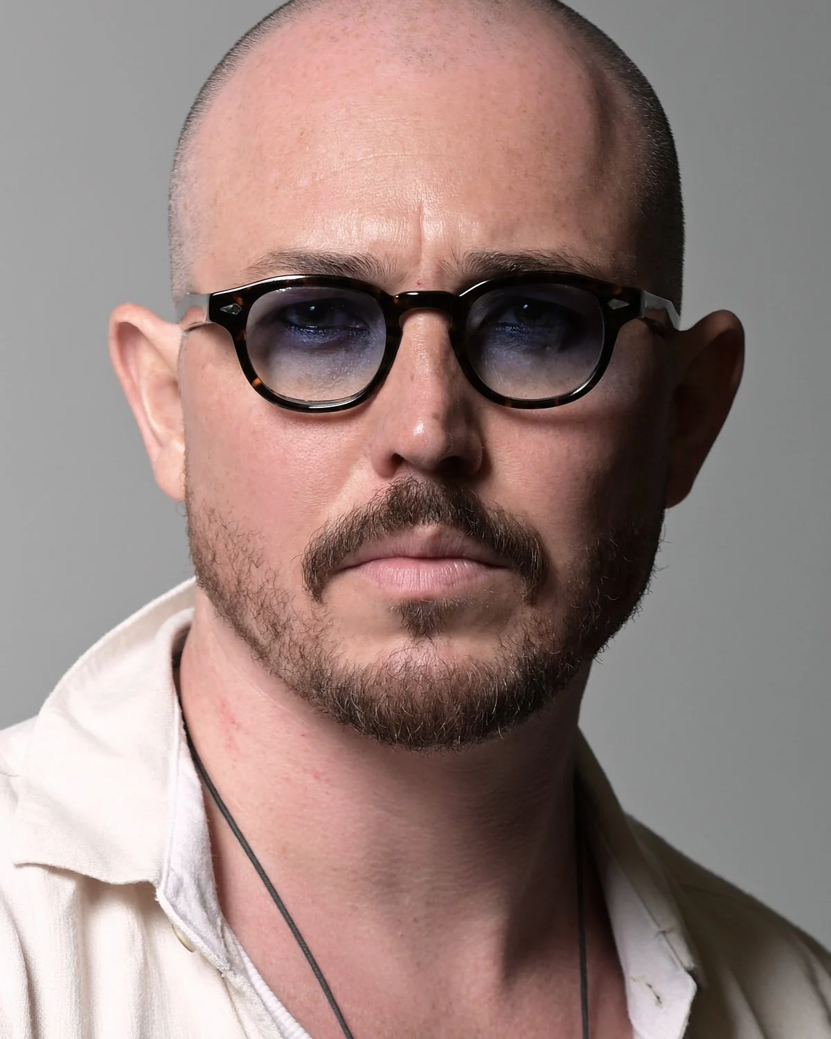 Close-up portrait of a man with a shaved head, glasses, and a beard, wearing a white shirt and a necklace, against a plain gray background.