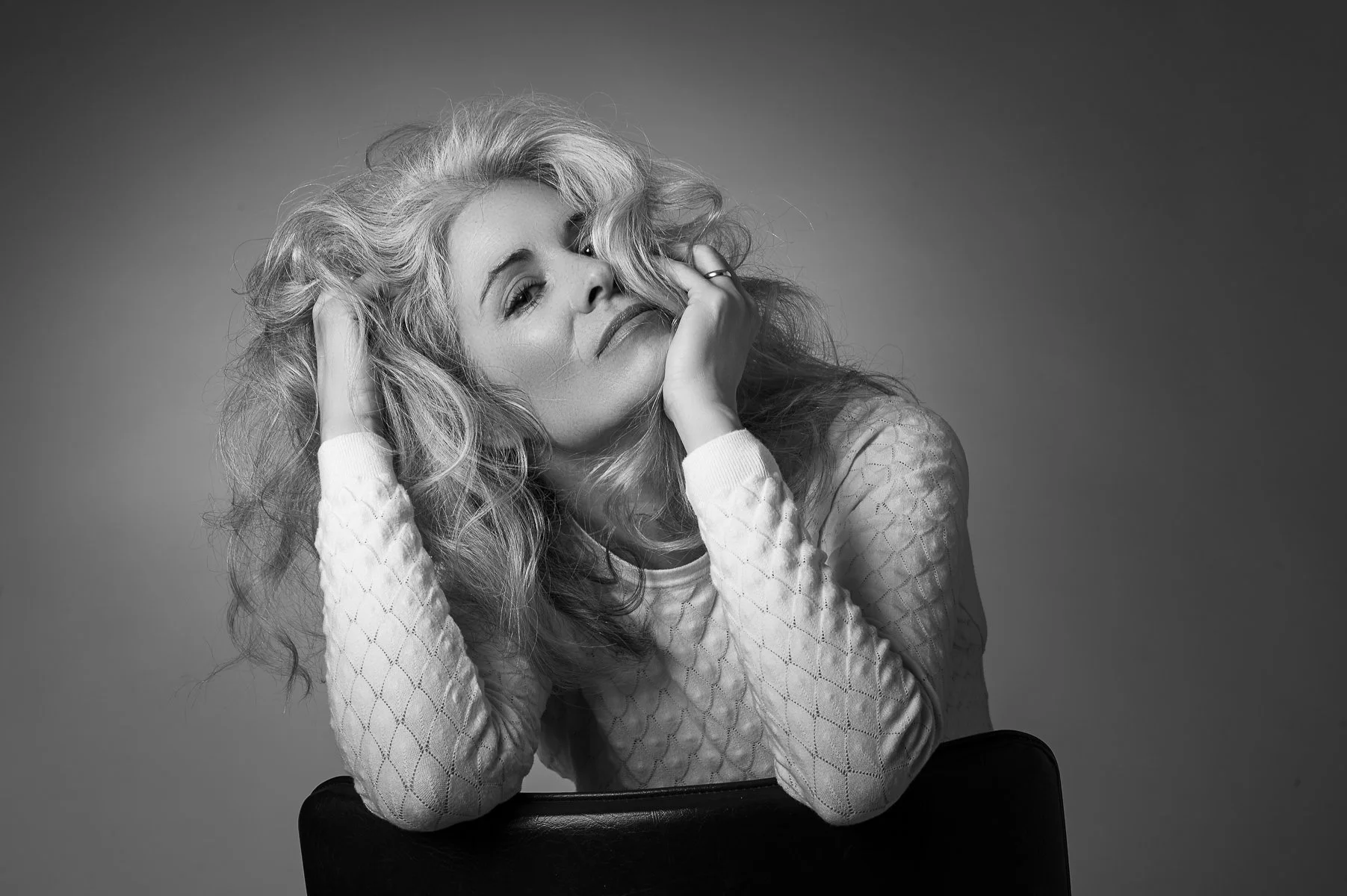 Black and white portrait of a woman in her thirties with wavy blonde hair, posing with her hand near her face, wearing a textured long-sleeve top, sitting on a chair against a plain background.