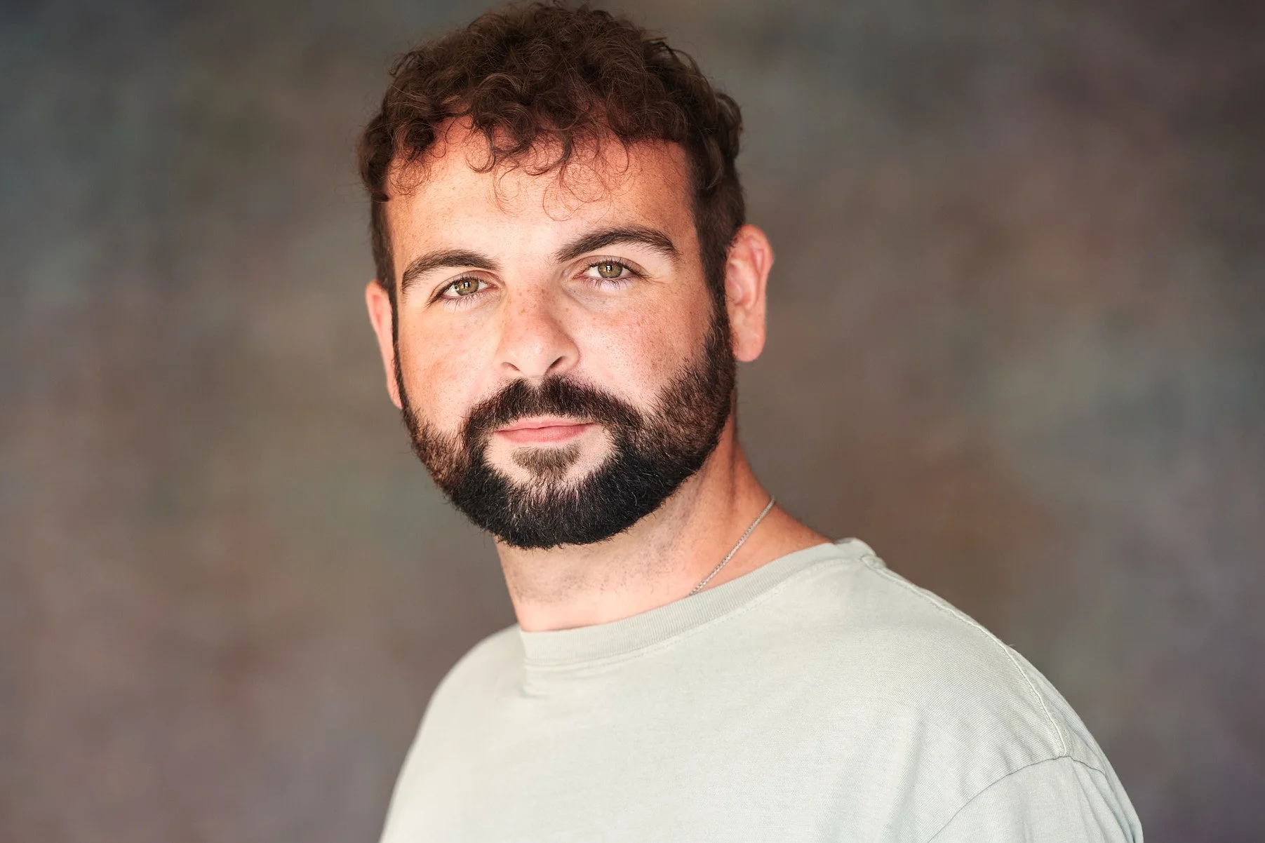 A portrait of a man with curly brown hair, a full beard, and hazel eyes, wearing a light gray shirt and a silver chain, against a softly blurred background.