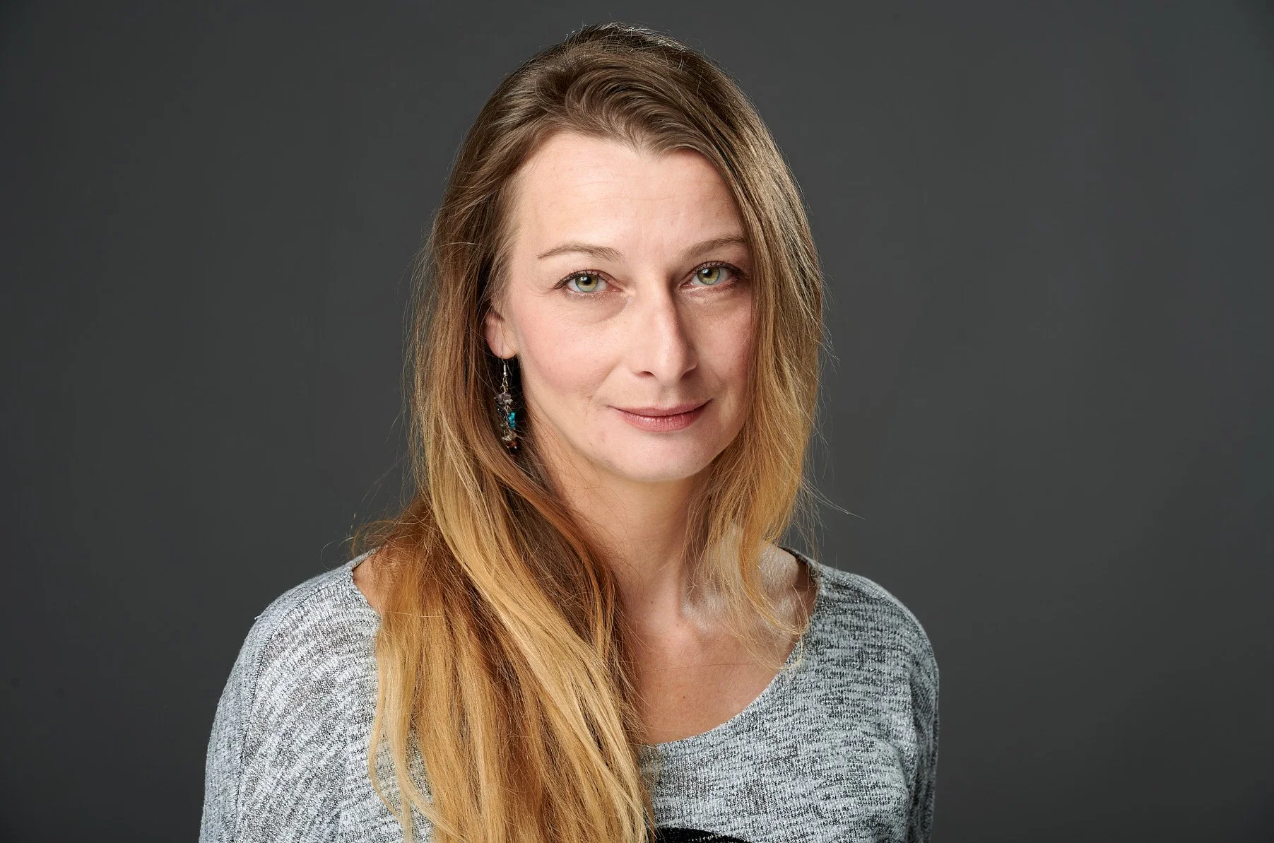 Portrait of a woman with long, light brown and blonde hair, wearing a gray top and colorful earrings, against a dark gray background.