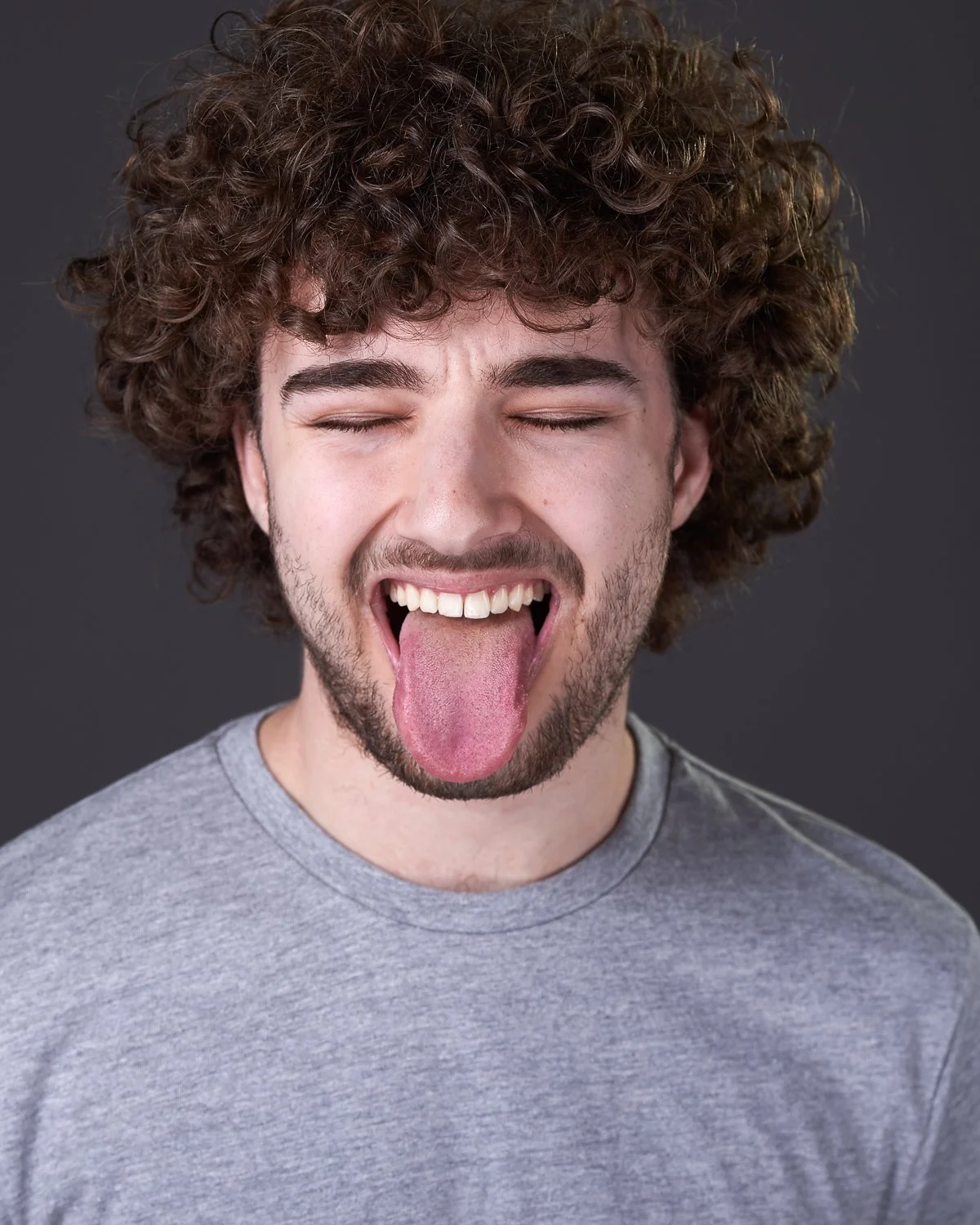A young man with curly brown hair sticking out his tongue and smiling with eyes closed, wearing a gray t-shirt against a dark background.
