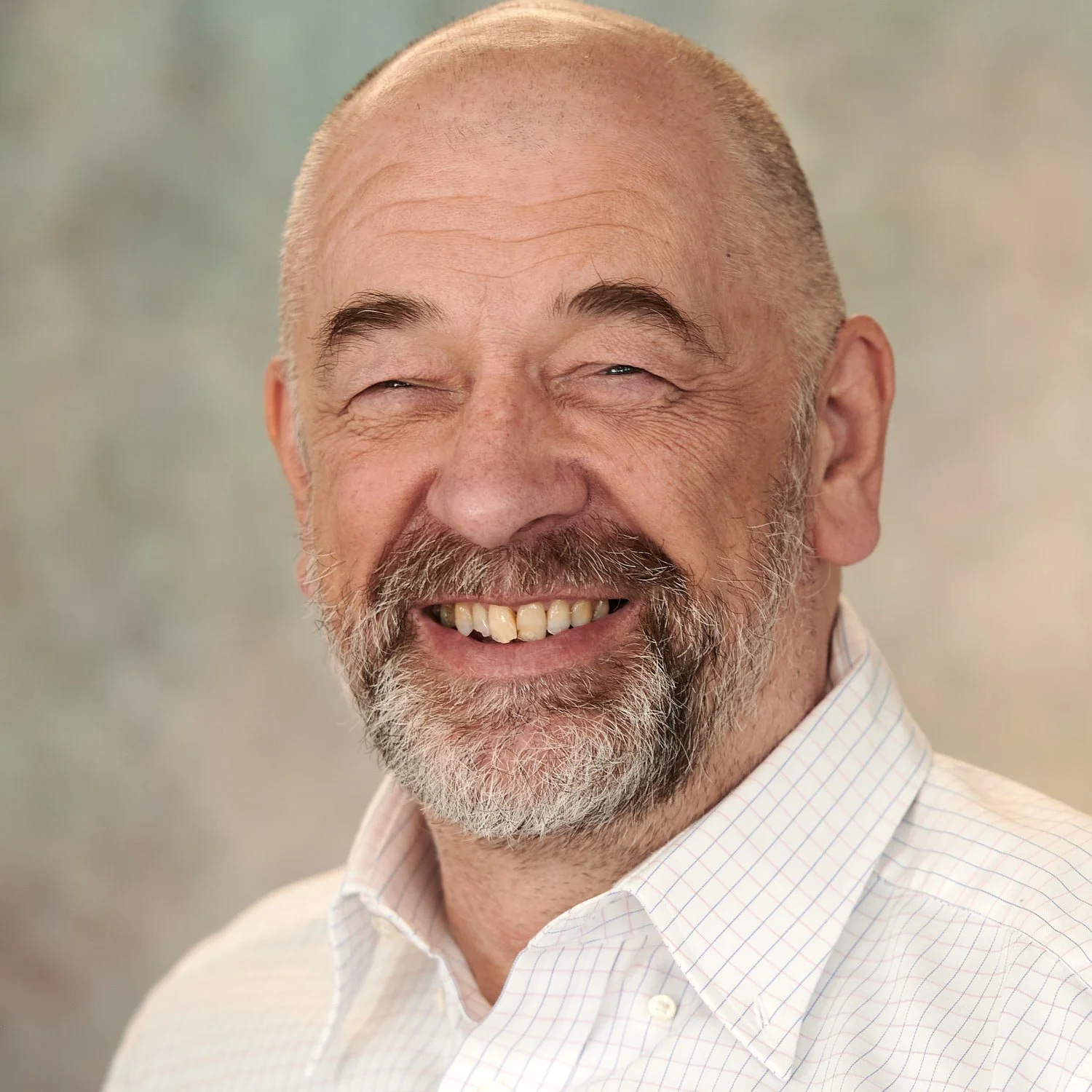 A smiling older man with a beard and mustache, wearing a light-colored, checkered button-up shirt, with a blurred neutral background.