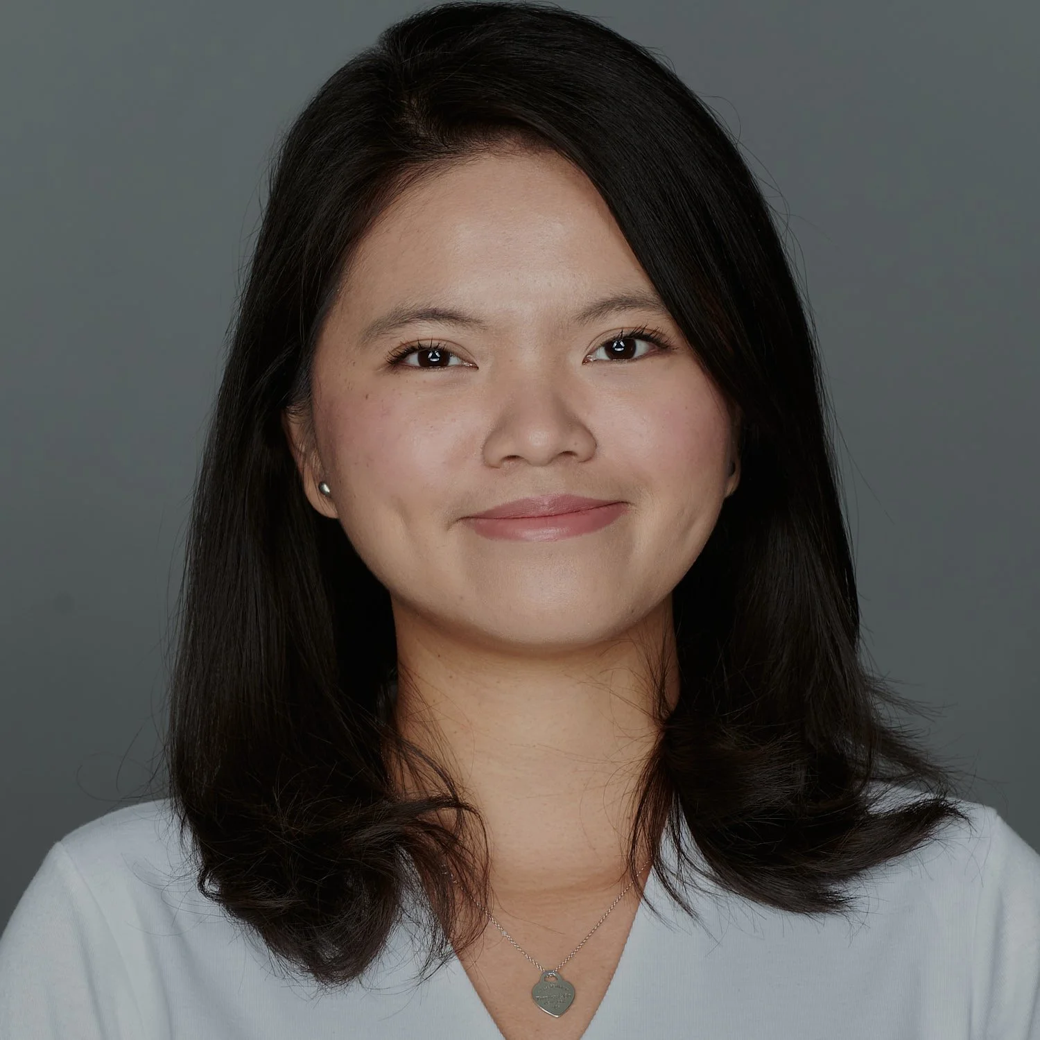 A woman with shoulder-length dark hair wearing earrings and a necklace, smiling against a gray background.