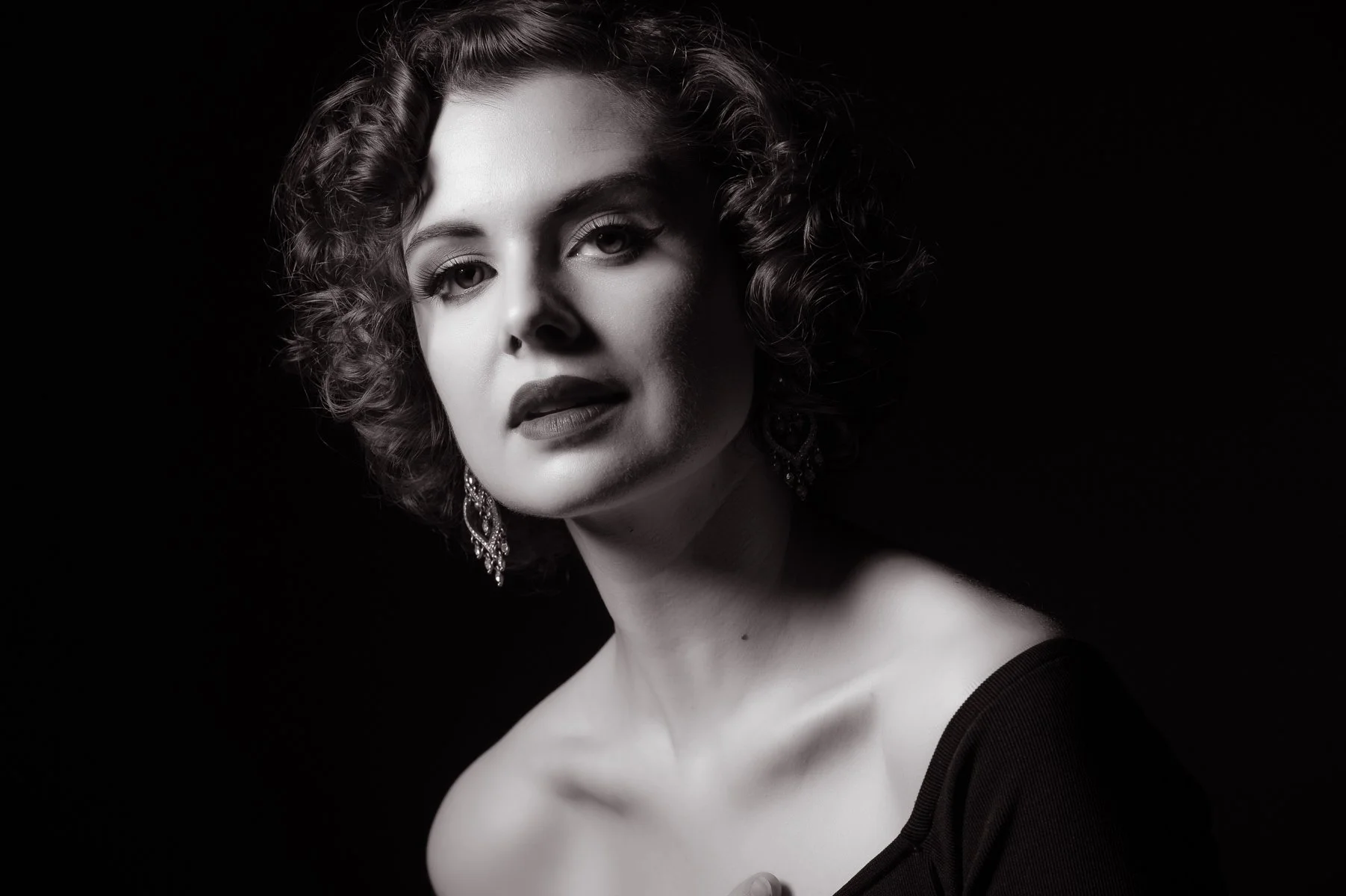 A black and white portrait of a woman with curly hair, wearing earrings and a dark top, against a dark background, photographed in the Hollywood style of the 1940s.