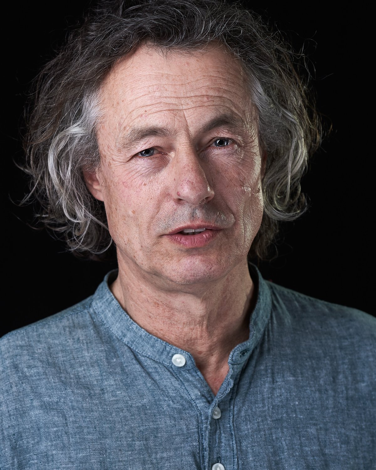 Close-up portrait of a mature man with gray, curly hair, wearing a light blue button-up shirt, against a black background.