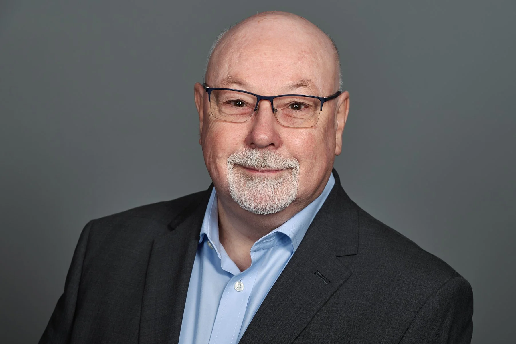 A headshot of a smiling, middle-aged man with glasses, a light blue shirt, and a dark gray suit against a gray background.