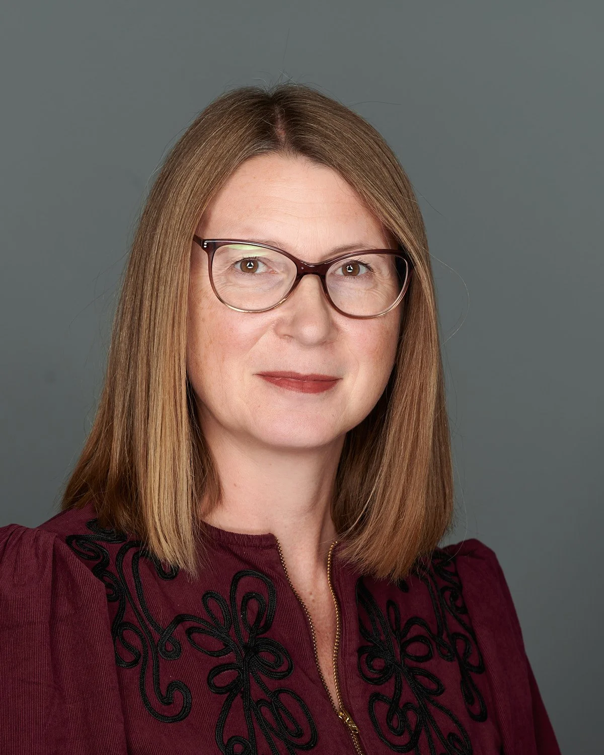 A woman with shoulder-length light brown hair, glasses, and a burgundy top with black embroidery, posing against a gray background.
