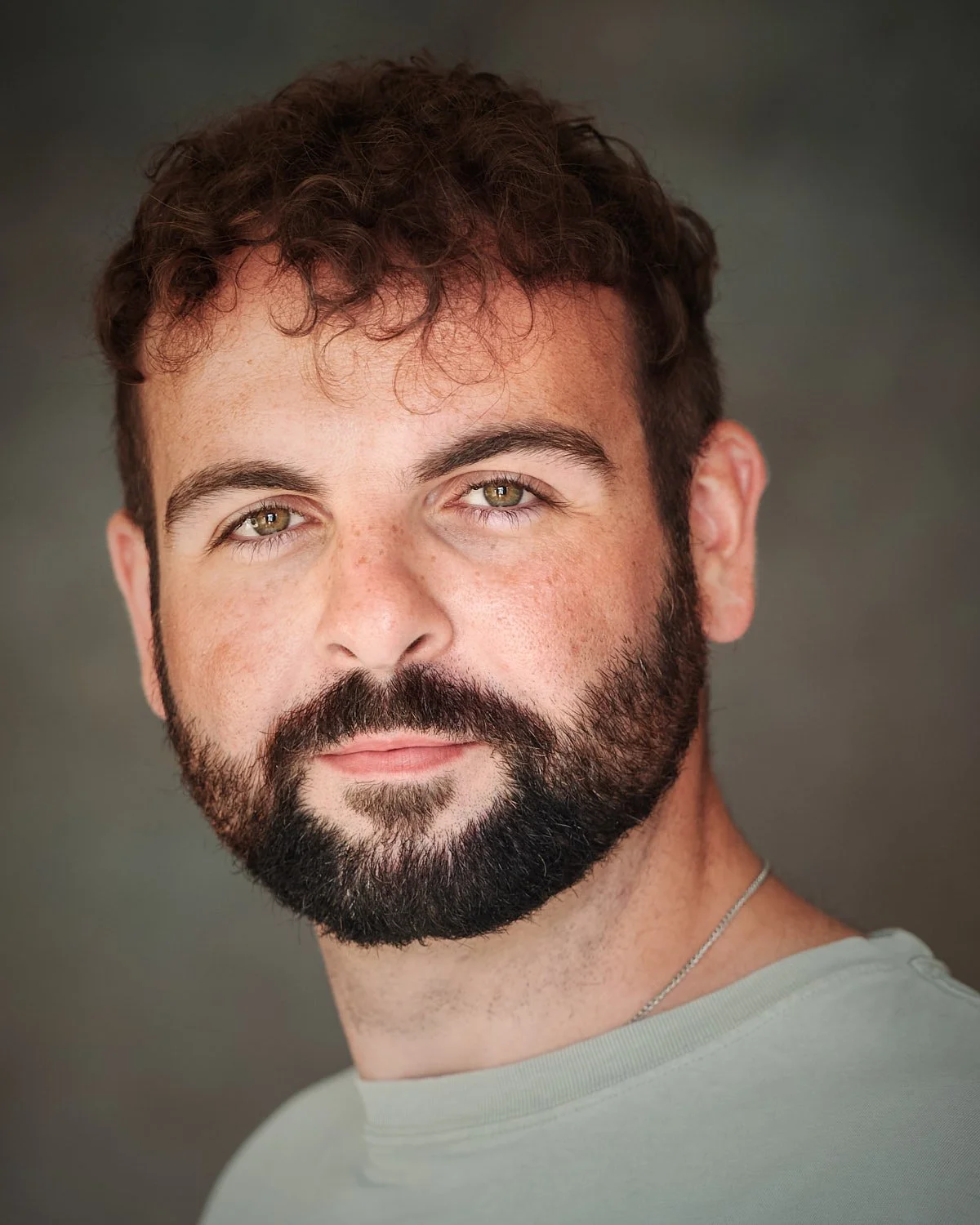 Close-up portrait of a man with curly brown hair, hazel eyes, and a full beard, wearing a light gray shirt and a silver chain necklace, against a blurred background.