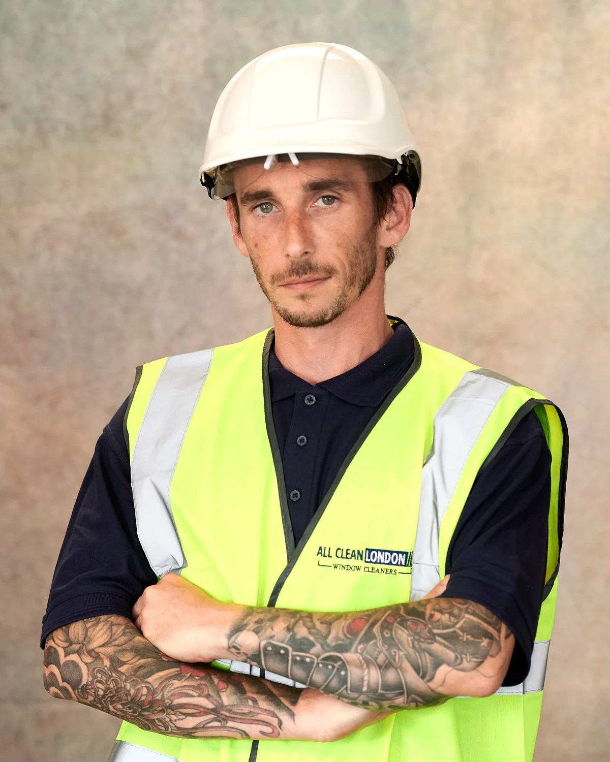 A young man with tattoos on his arms, wearing a white safety helmet and a reflective yellow safety vest with a company logo, standing with arms crossed against a neutral background.