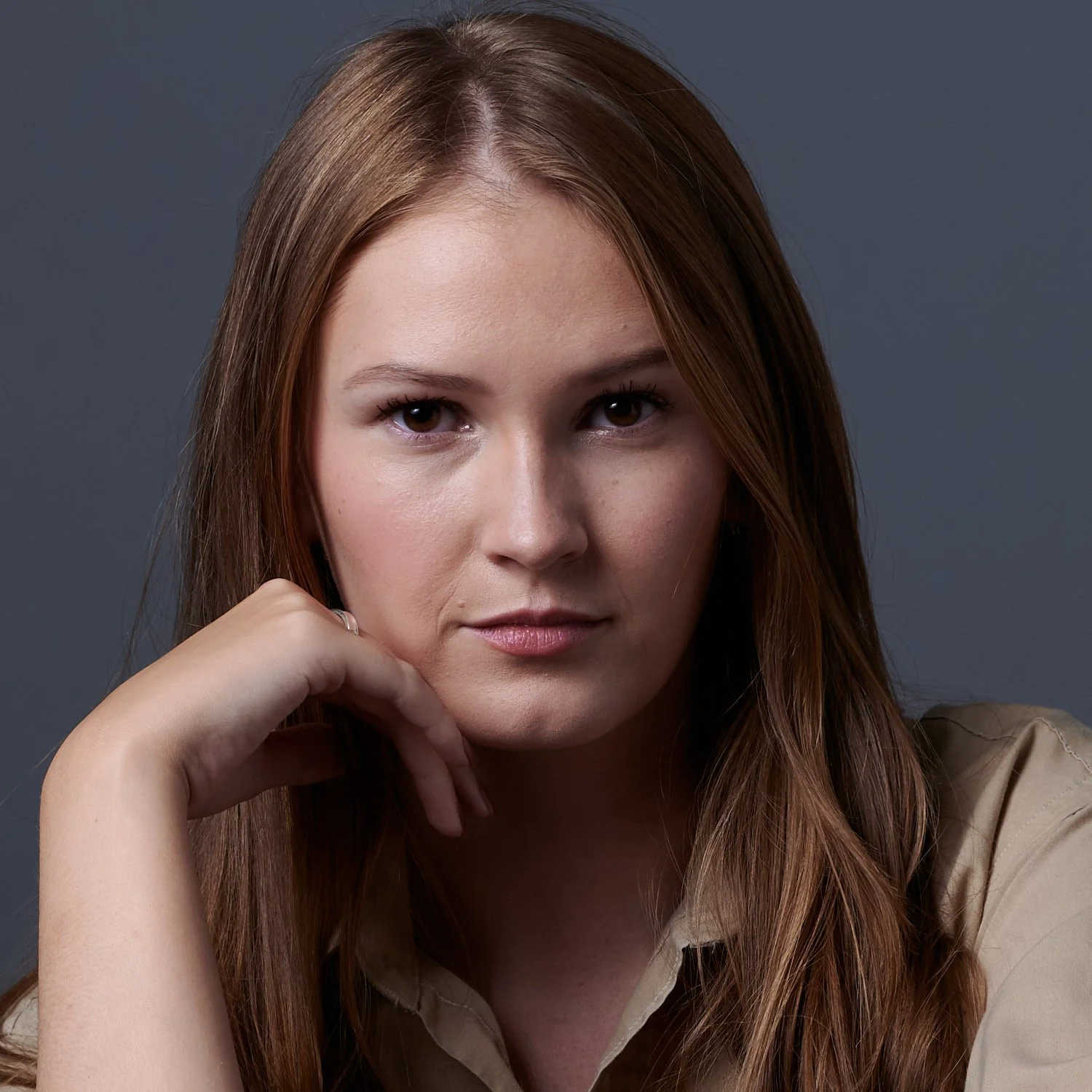 Close-up portrait of a woman with long red hair, looking directly at the camera with a neutral expression, resting her chin on her hand against a gray backdrop.
