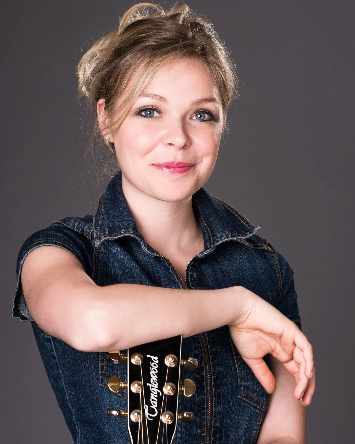 A woman with blonde hair styled in a loose updo, wearing a sleeveless denim shirt, poses with her arm resting on her shoulder and smiling at the camera. She holds a black guitar headstock with gold tuning pegs and the word 'Wannabegood' written on it