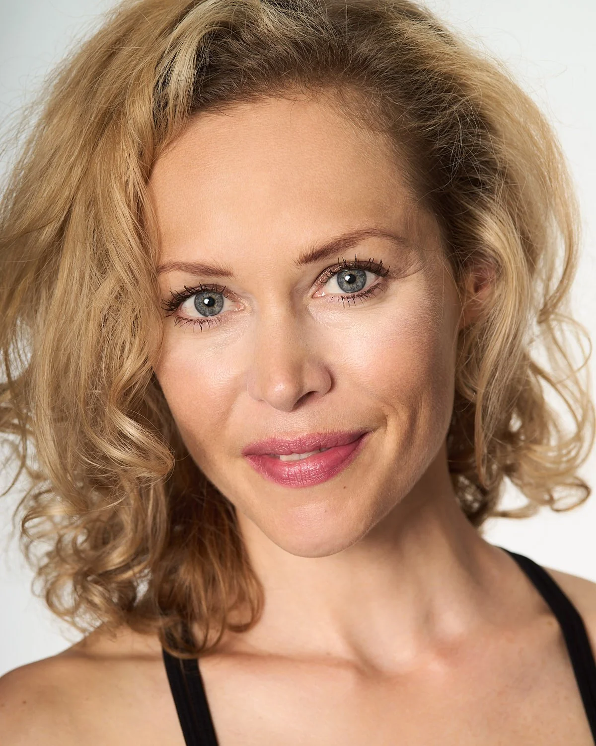 A close-up portrait of a woman with wavy blonde hair and blue eyes, smiling slightly, wearing a black top, against a neutral background.