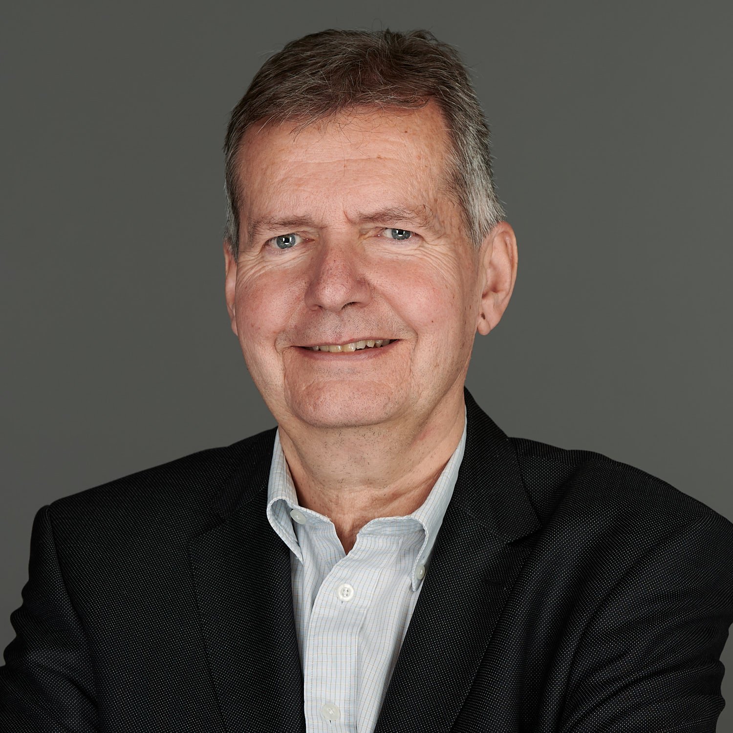 A middle-aged man with short gray hair, blue eyes, and a friendly smile, wearing a dark suit jacket and a white collared shirt, posing against a plain gray background.