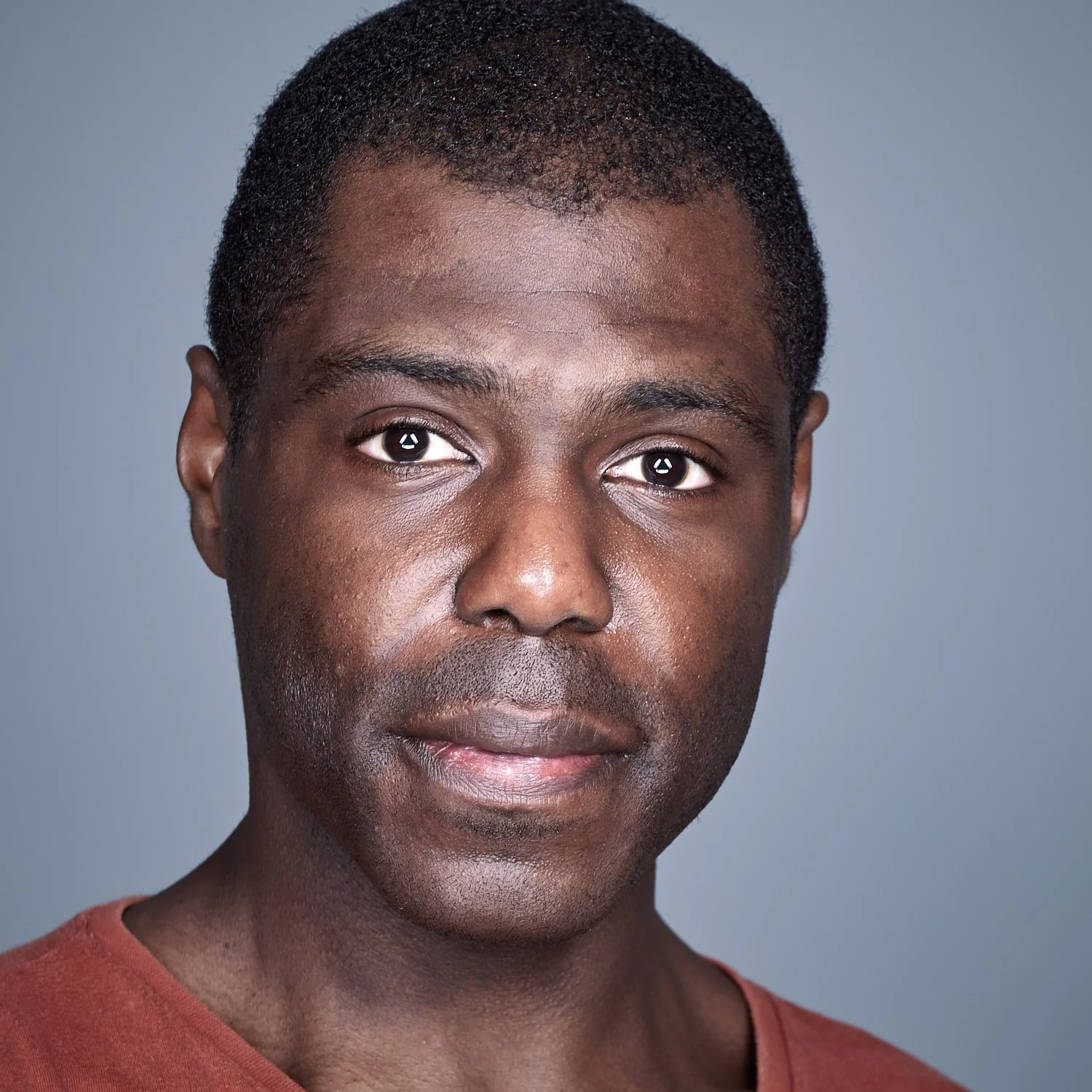 A close-up portrait of a young African American man with short, curly hair, wearing a reddish-brown shirt against a neutral gray background.