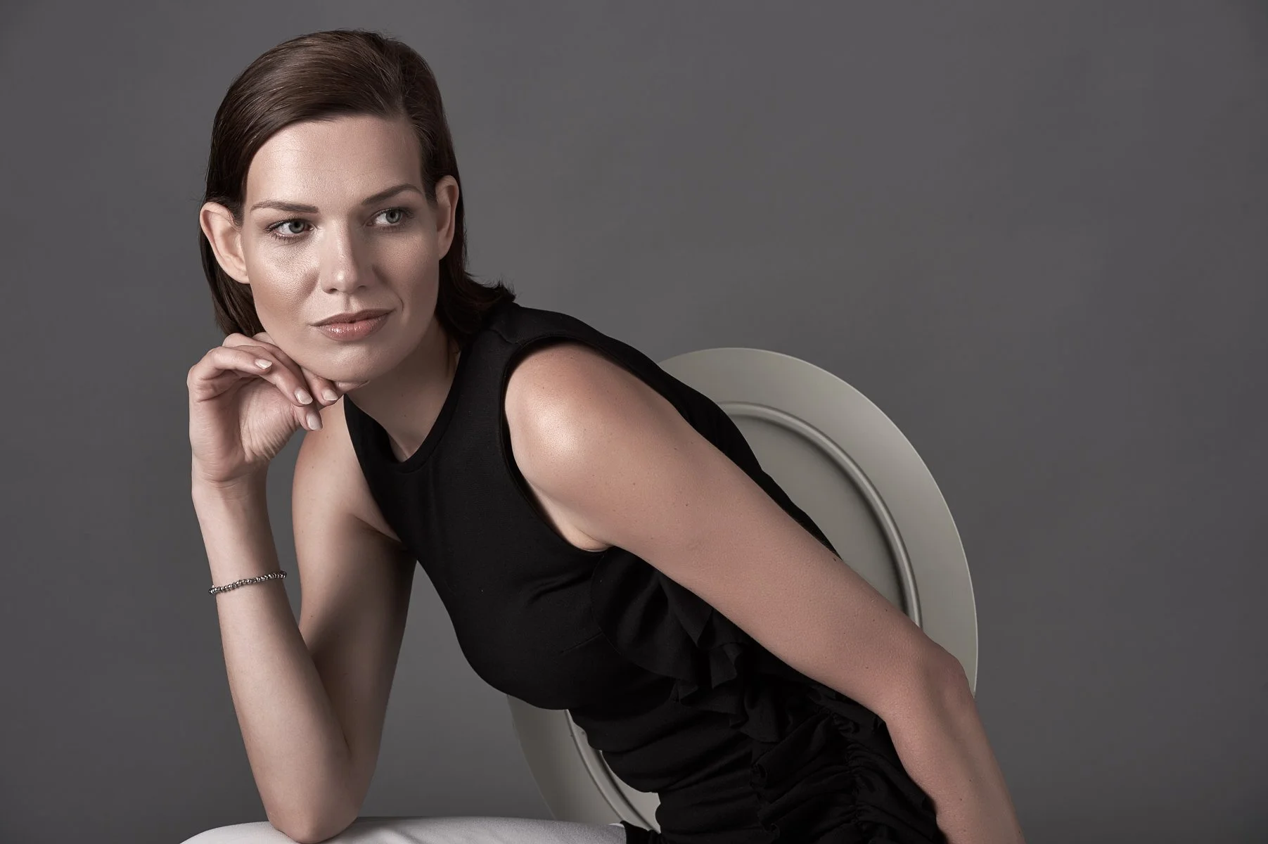 A young woman with short brown hair wearing a sleeveless black dress, seated on a white chair with a gray background, resting her chin on her hand and looking slightly to her right.