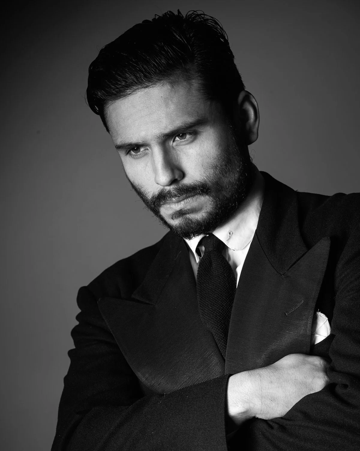 Black and white portrait of a man with a beard, wearing a suit and tie, looking down thoughtfully with arms crossed.