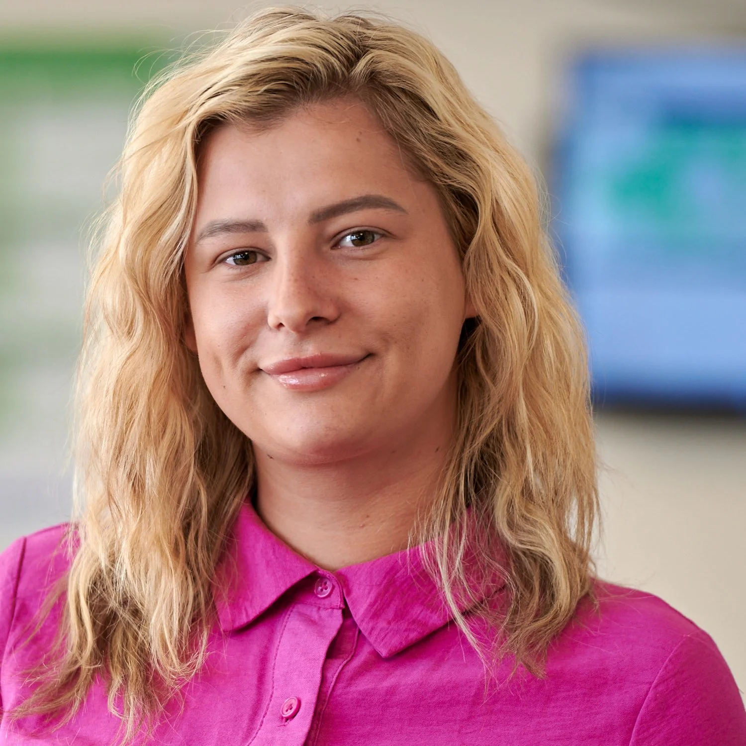 A young woman with blonde, wavy hair wearing a bright pink collared shirt, smiling softly in an indoor setting with a blurred background.