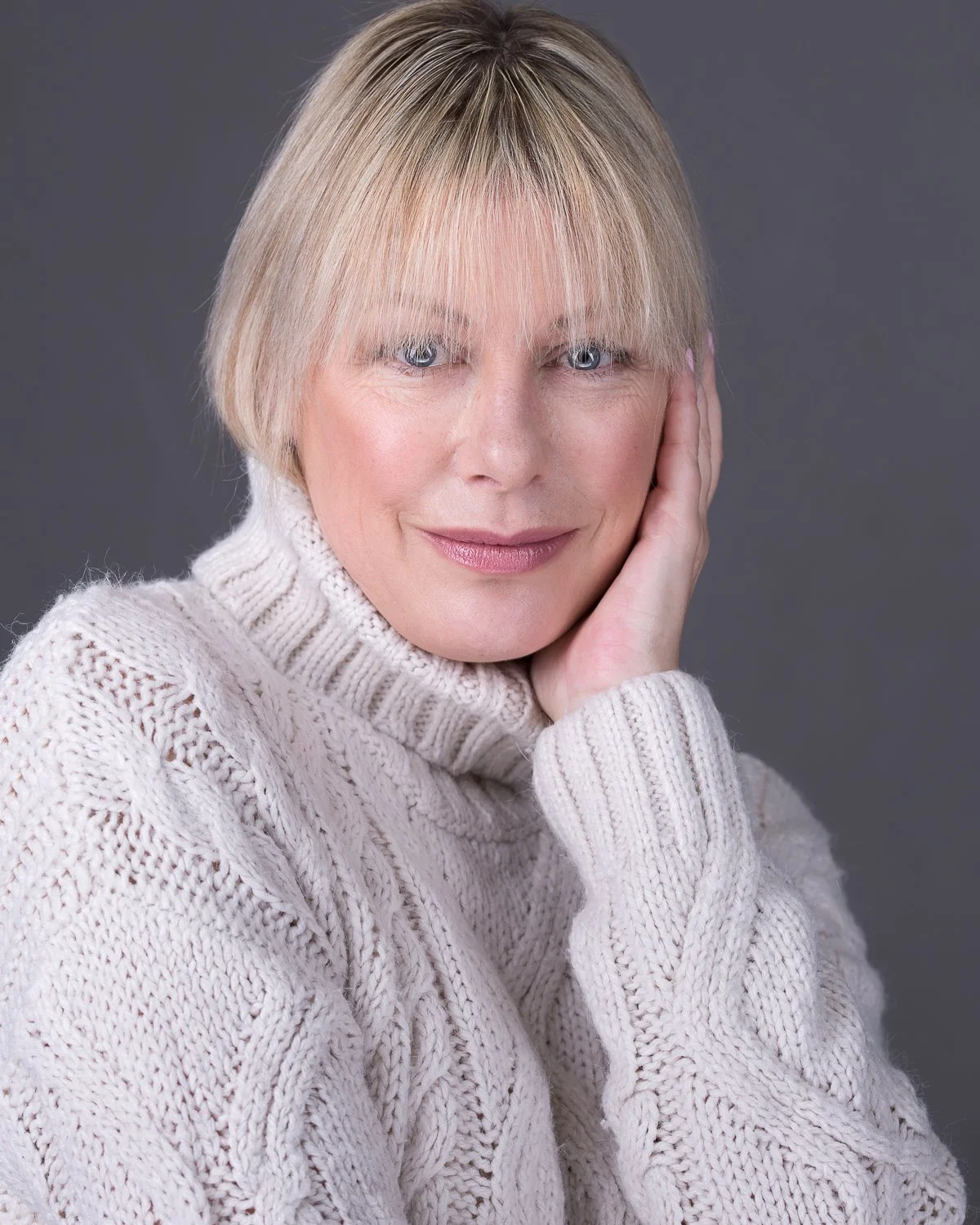A woman with blonde hair and blue eyes, wearing a cream-colored knit sweater, posing with her hand on her face against a grey background.