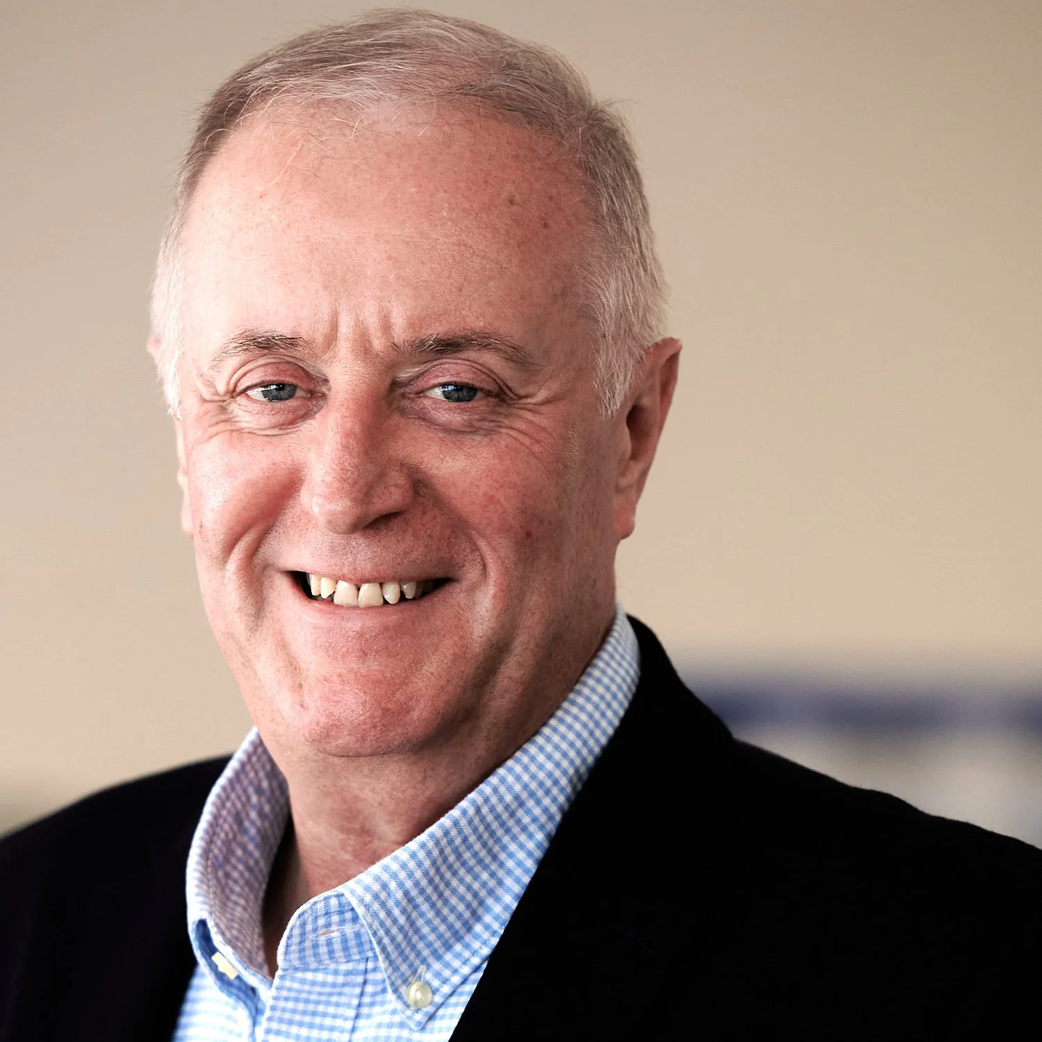 A middle-aged man with short, grey hair, blue eyes, and a wide smile showing his teeth. He is wearing a blue and white checkered shirt and a dark blazer. The background is plain and light-colored.