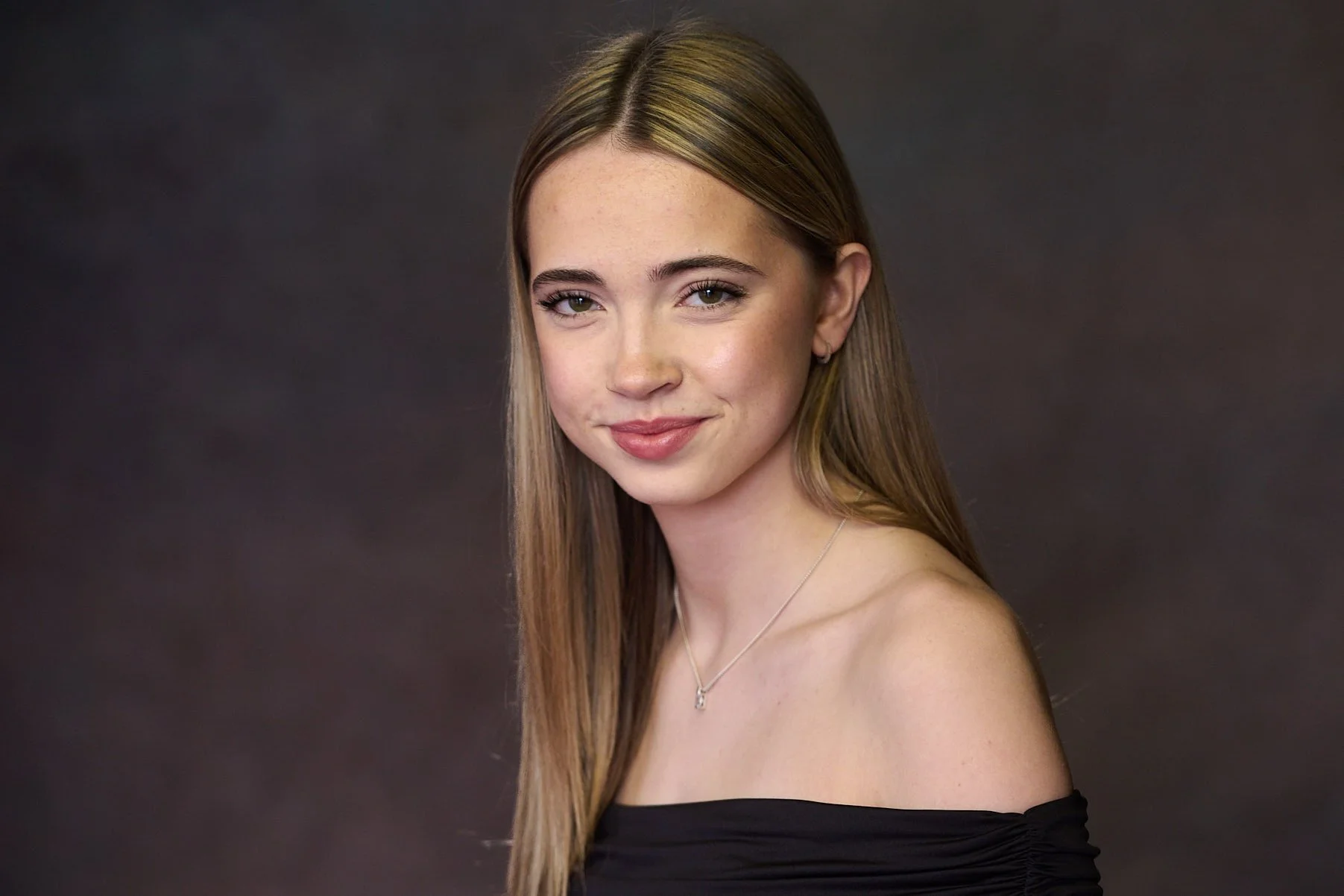 Young woman with long, straight blonde hair, wearing an off-the-shoulder black top, posing against a dark background.