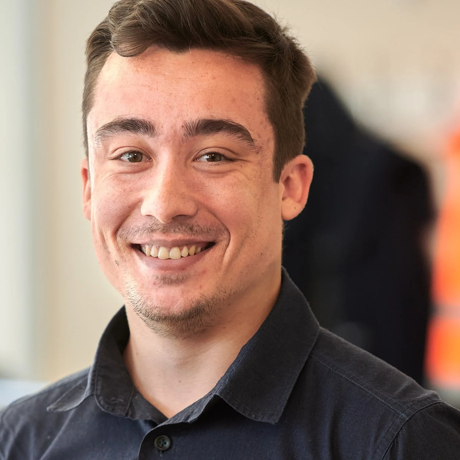 Close-up of a young man smiling, wearing a black collared shirt, with short brown hair and a light beard, indoors with a blurred background.