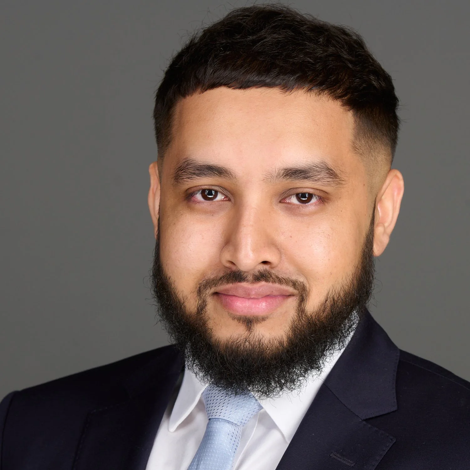 Professional headshot of a man in a dark suit, white shirt, and light gray tie, with a neutral gray background.