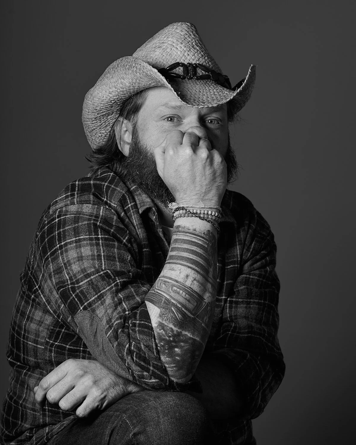 A man with a beard wearing a cowboy hat and plaid shirt, sitting with one hand covering his mouth and the other resting on his arm, in black and white studio portrait.