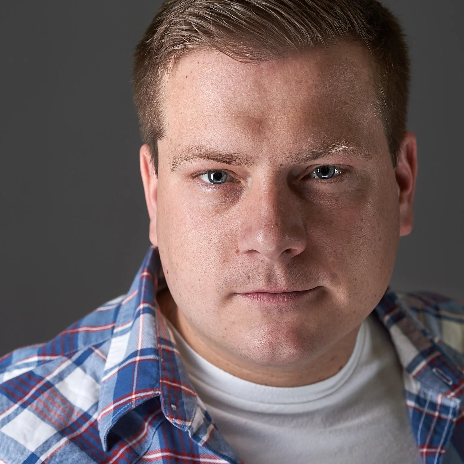 Close-up portrait of a young man with light skin, blue eyes, short brown hair, wearing a white t-shirt and a blue plaid shirt, against a dark gray background.